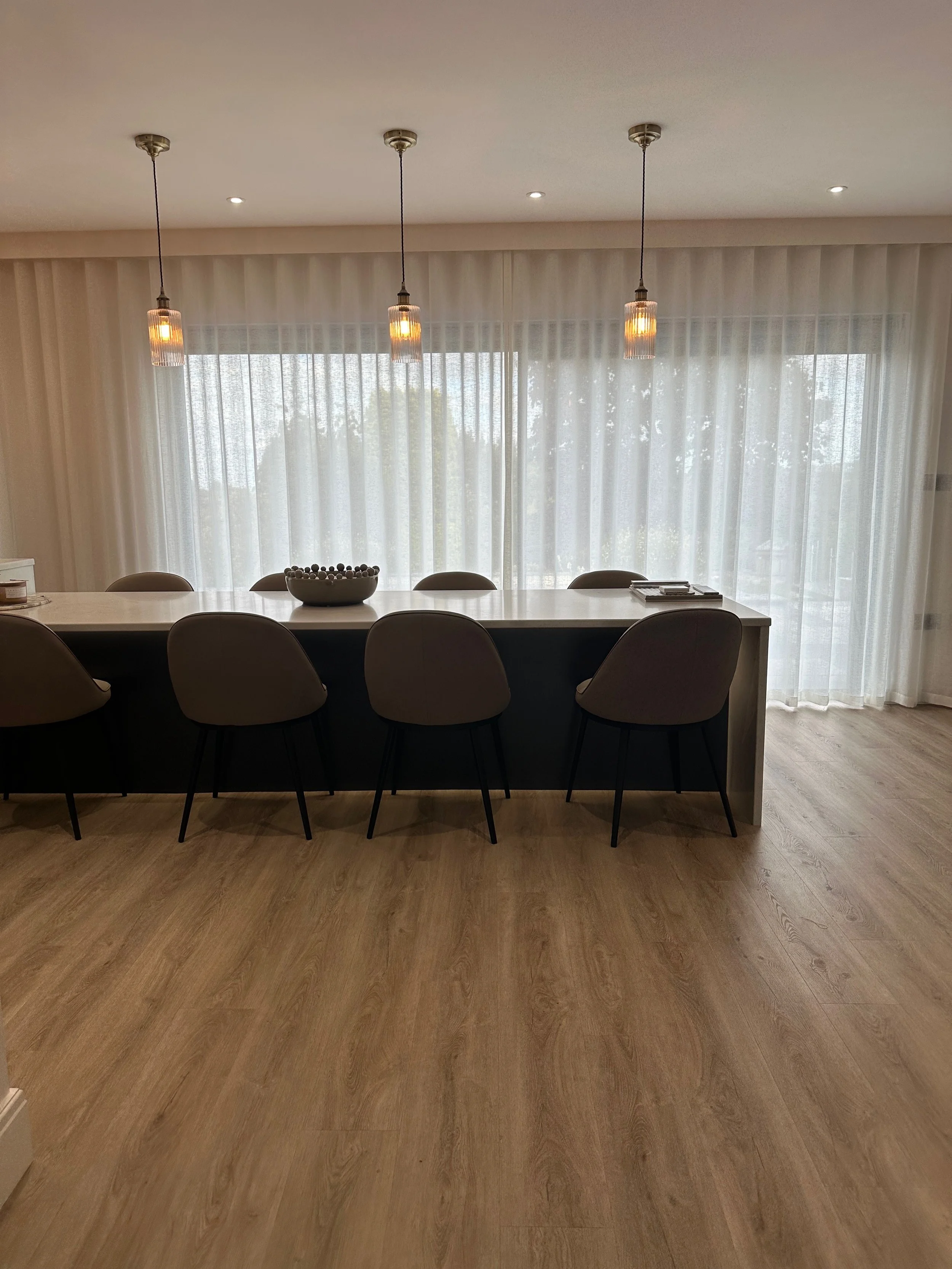 Modern dining room with a long white table, six beige chairs, pendant lights, sheer white curtains, hardwood flooring, and a large window showing trees outside.