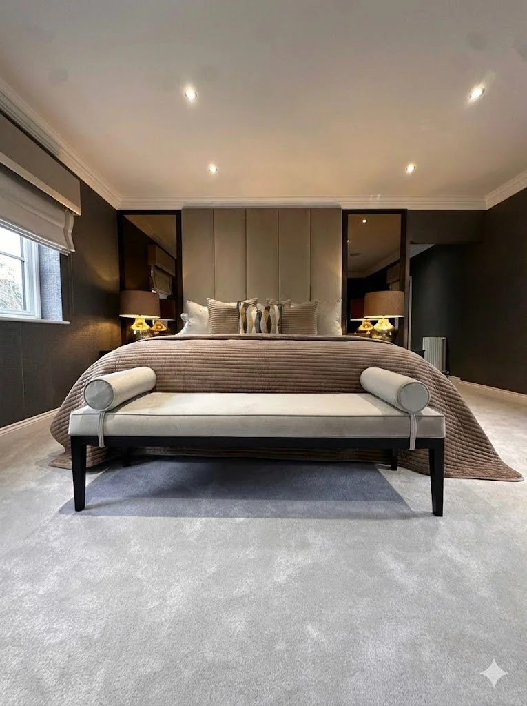 Modern bedroom with a large bed, beige upholstered headboard, decorative pillows, two nightstands with lamps, a bench at the foot of the bed, window with a blinds, and dark walls with recessed ceiling lights.