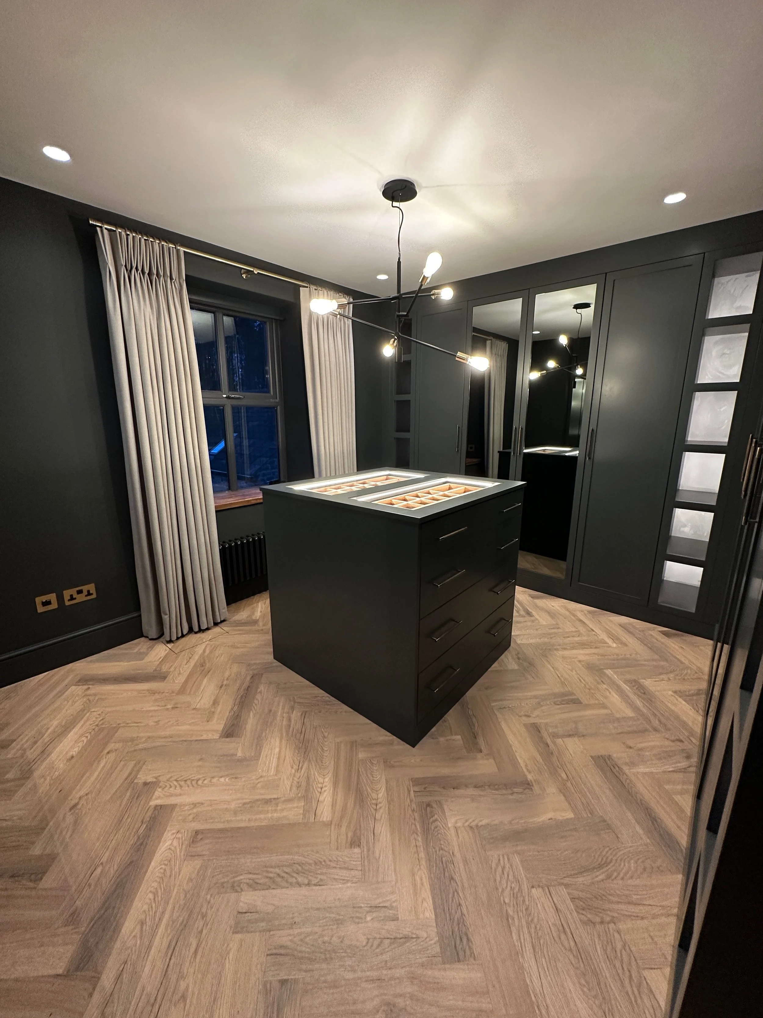 A modern kitchen with dark cabinets and a black island, herringbone wooden flooring, a large window with curtains, and a contemporary chandelier with multiple bulbs.