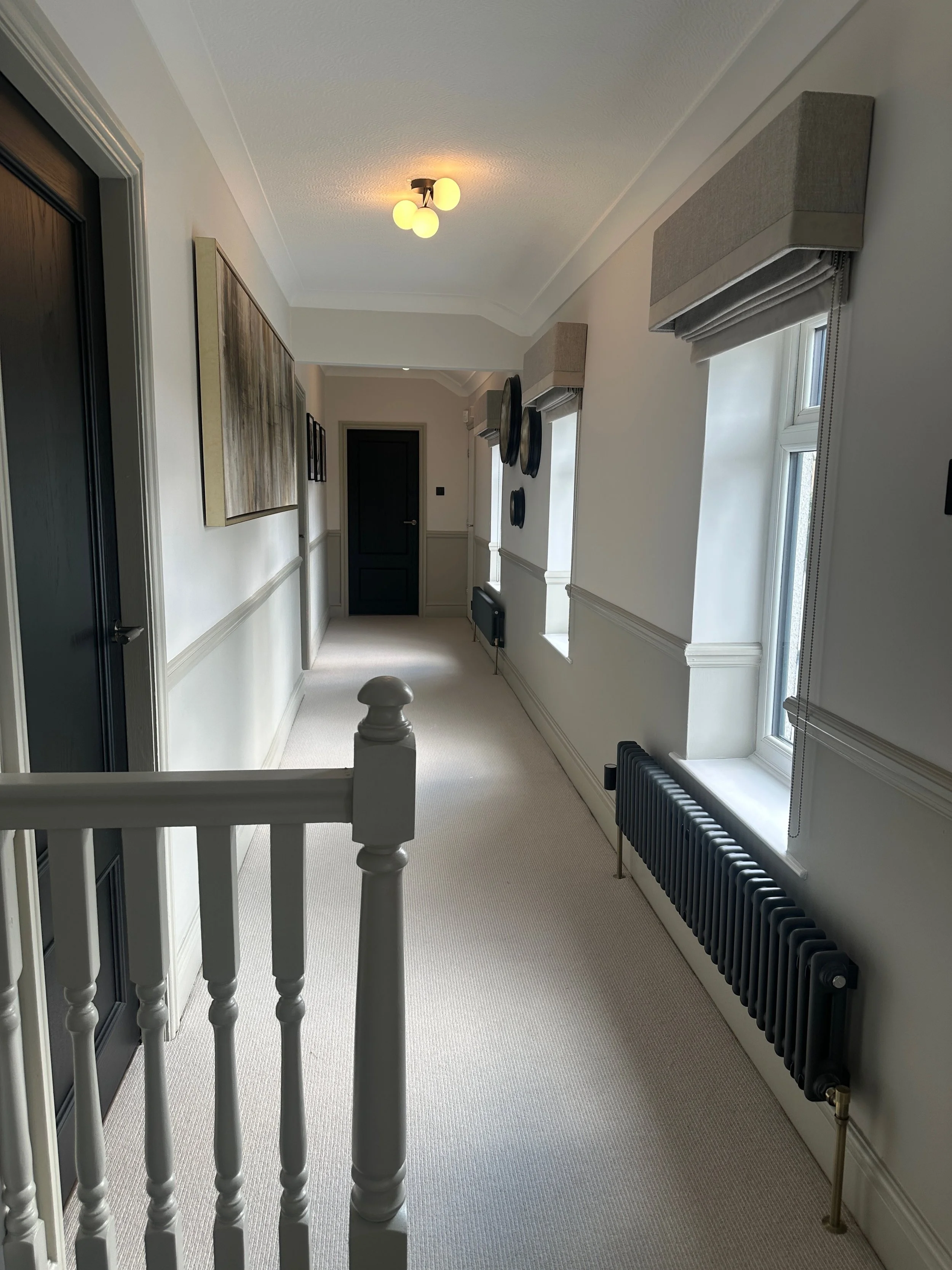 Bright hallway with two windows with beige valances, black radiators, white walls with wainscoting, artwork on the wall, and a ceiling light fixture with three bulbs.