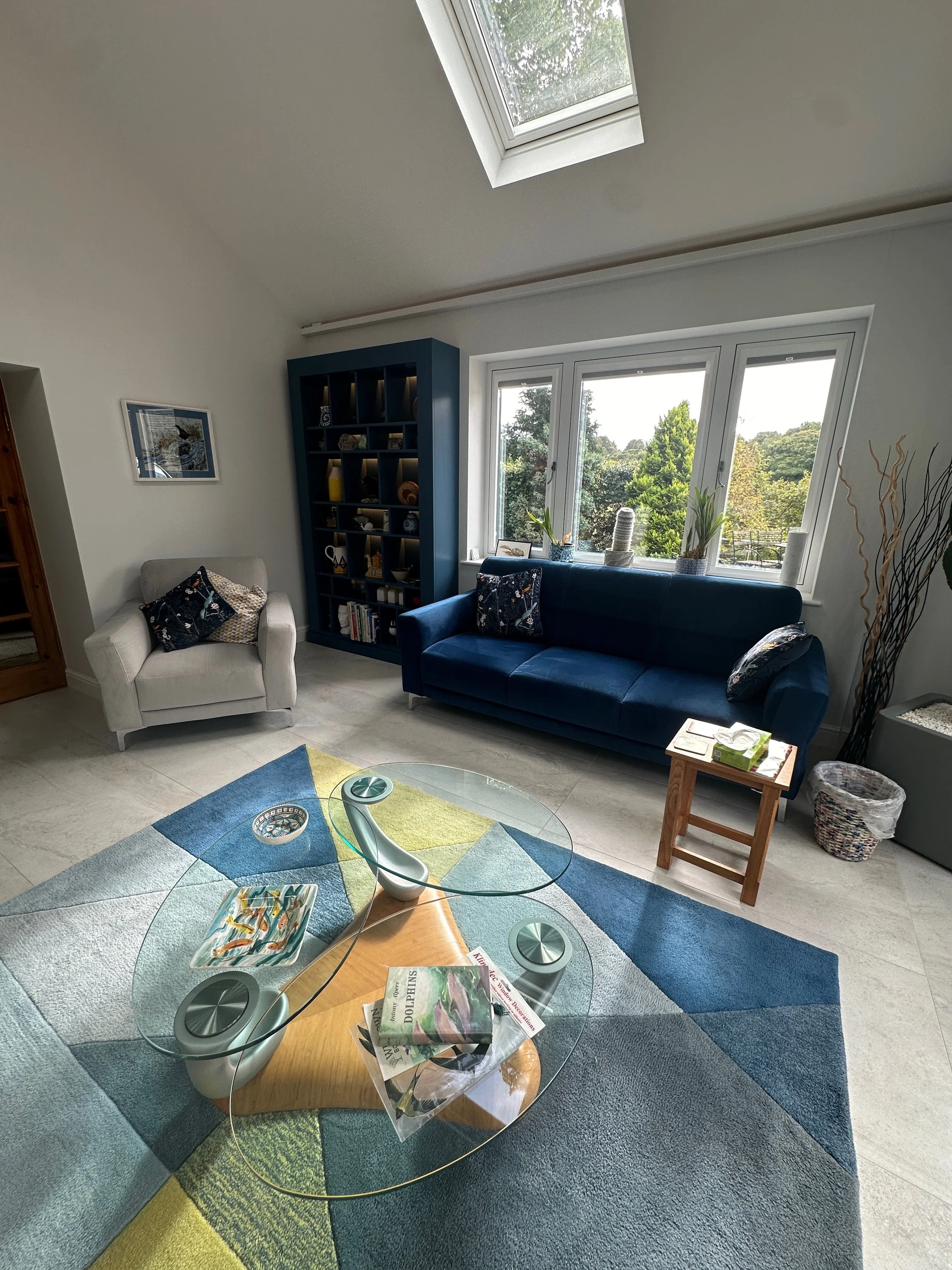 Living room with a white armchair, a dark blue sofa, a colorful rug, a glass coffee table, a tall blue bookshelf, and a window with a scenic view of trees.
