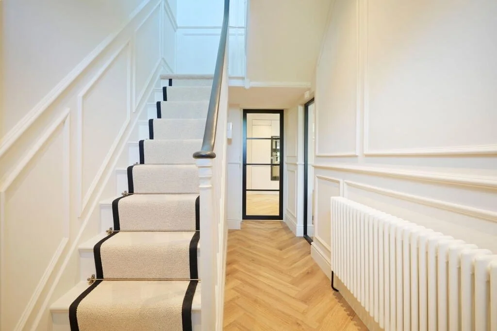 Interior view of a hallway and staircase with a beige runner rug, white walls with decorative molding, a white radiator, and a black framed glass door at the end.