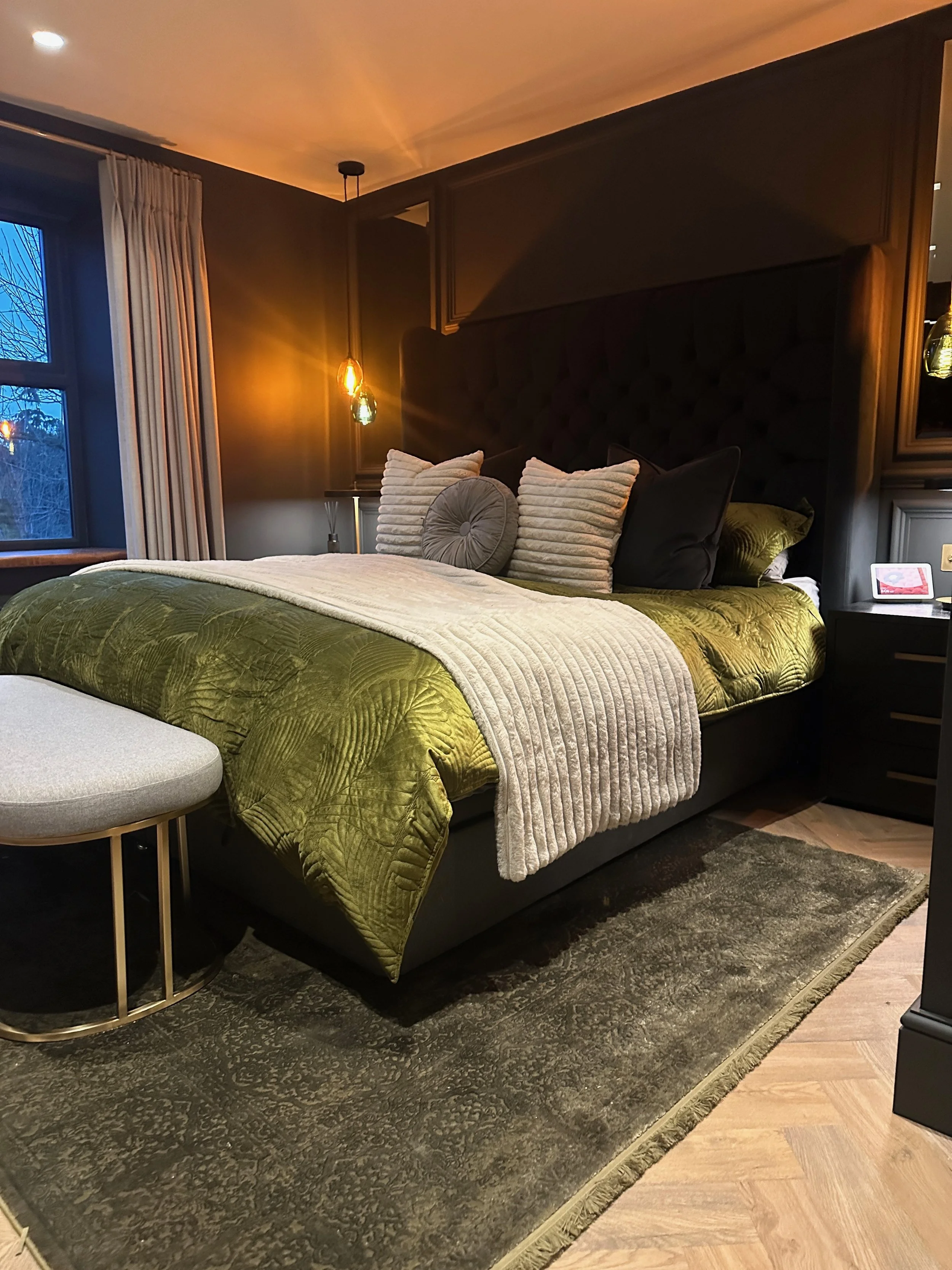 A neatly made bed with an olive green quilt, surrounded by multiple pillows including beige, black, and round gray ones. The bed has a tall, dark, tufted headboard, in a bedroom with dark walls, wooden flooring, and a dark rug. Two pendant lights hang on either side of the bed. There is a window with white curtains and a small black nightstand with a digital photo frame on it.