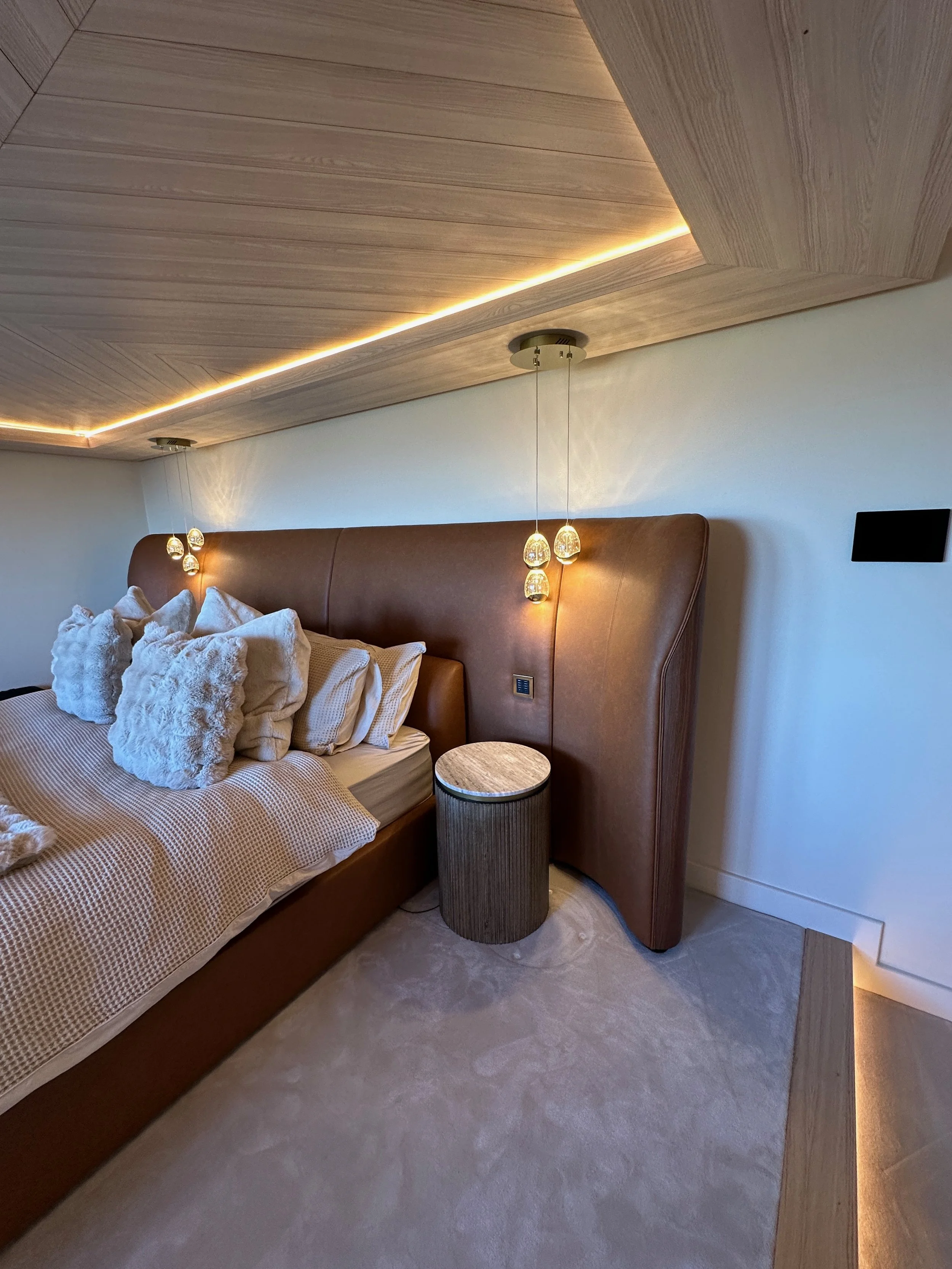 Close-up of a bedroom corner with a bed featuring multiple pillows, a round side table, and wall-mounted pendant lights with warm lighting, a wood-paneled ceiling, and a neutral-colored carpet.