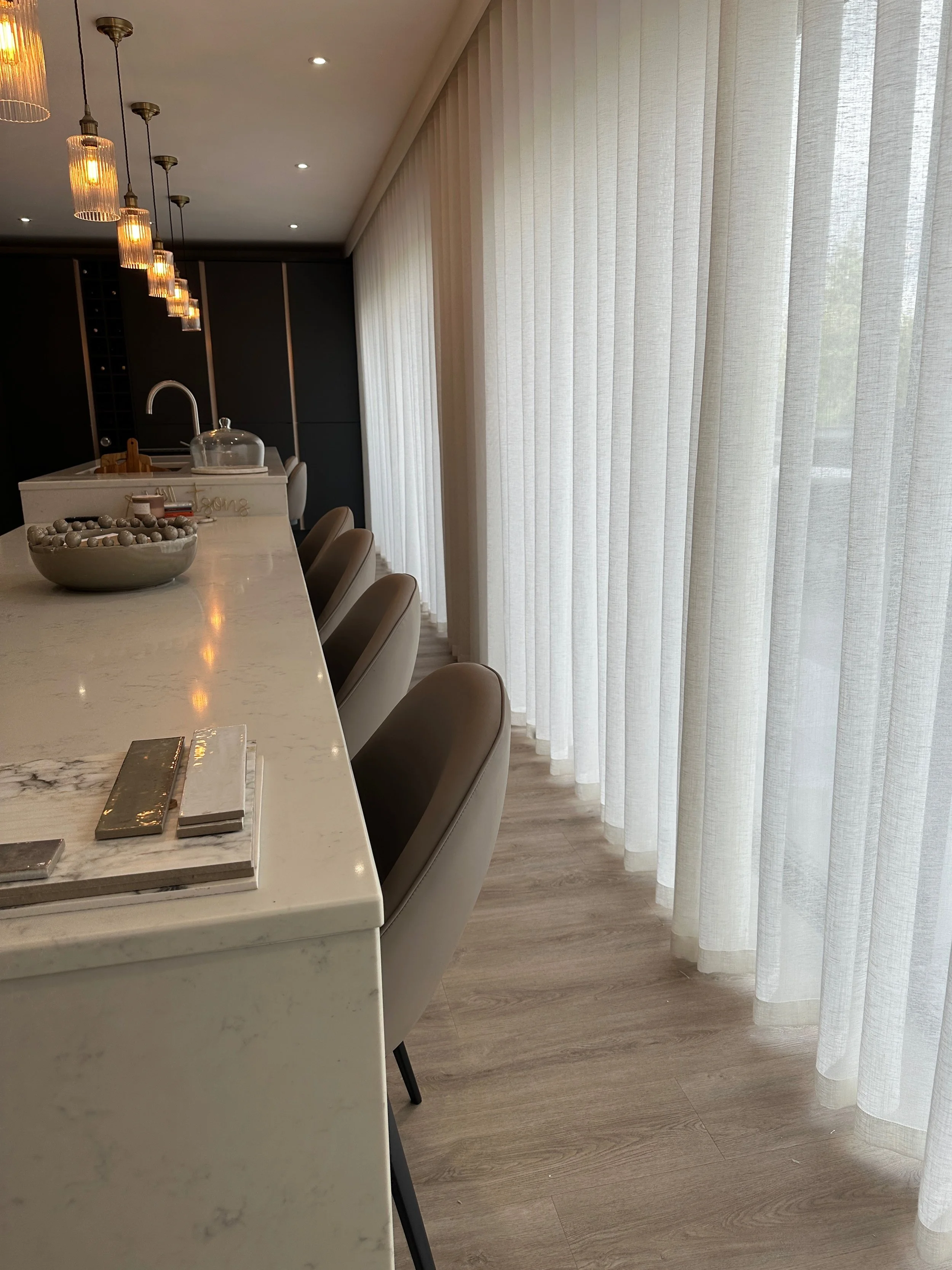 Modern kitchen with a white marble island, beige chairs, and sheer curtains blocking the view outside.