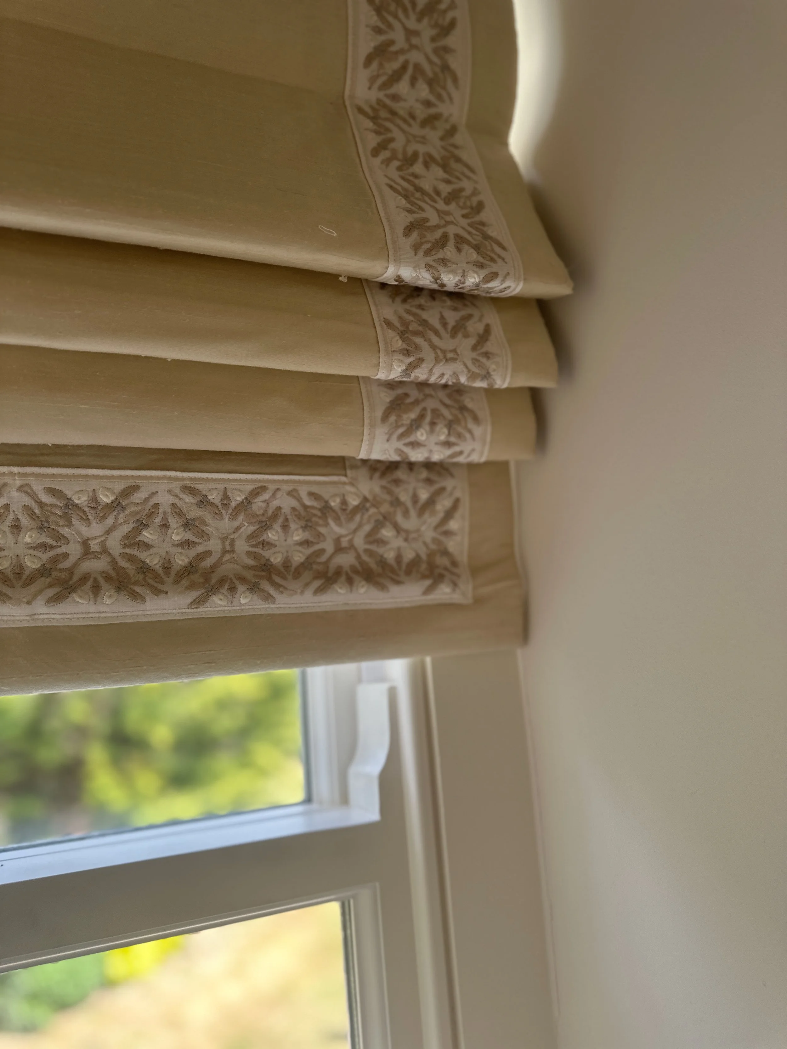 Close-up of a beige curtain with embroidered floral patterns, hanging in a room with a window showing greenery outside.
