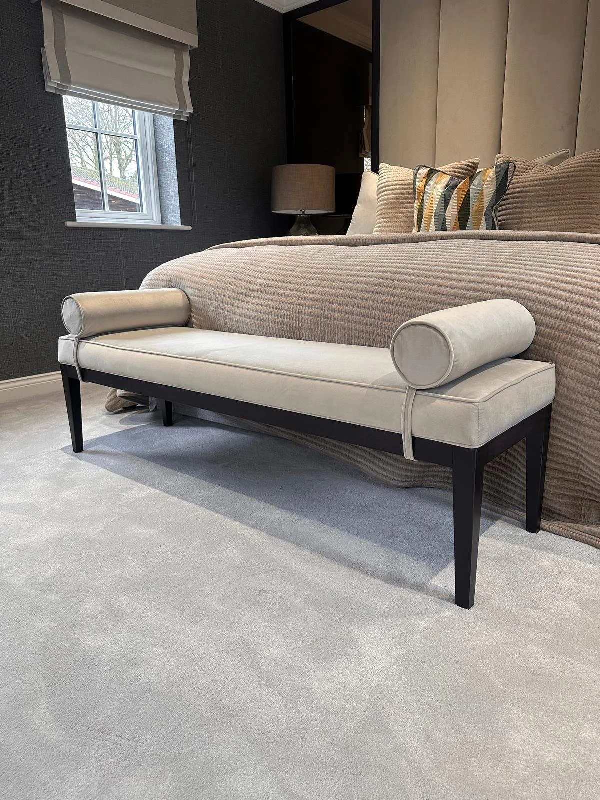 A living room with a bed, decorative pillows, a side table with a lamp, a window with blinds, and a cushioned bench at the foot of the bed.