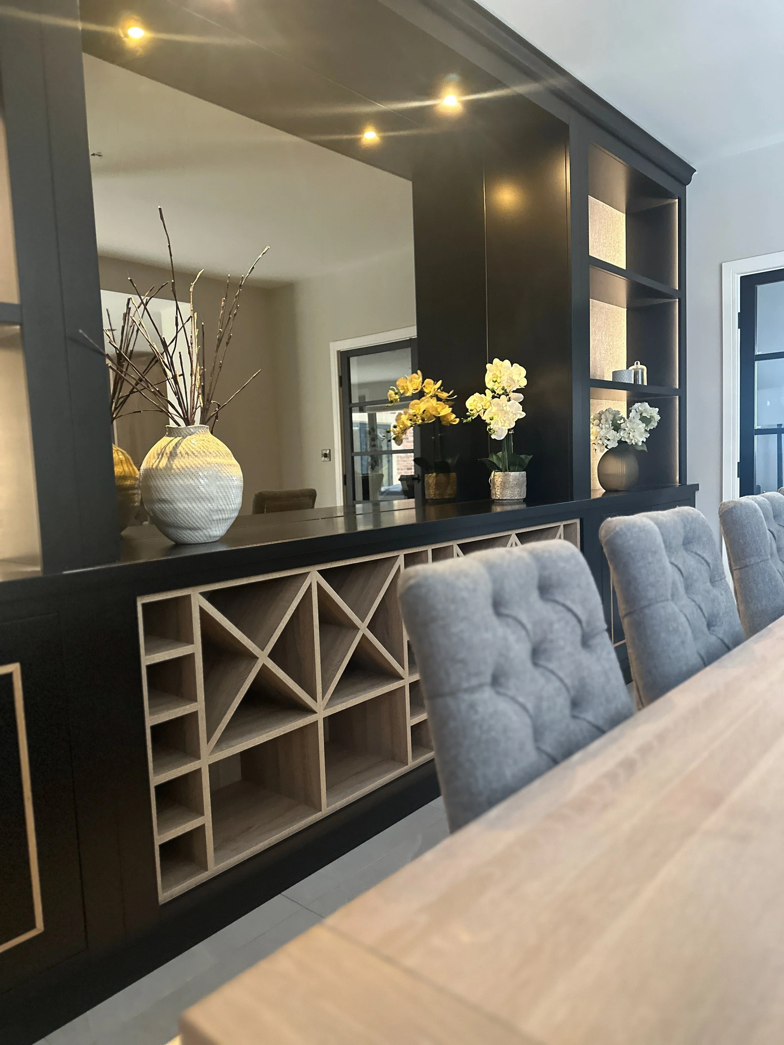 A dining area with a wooden table, gray upholstered chairs, and a black and wood shelving unit with decorative vases and flowers.