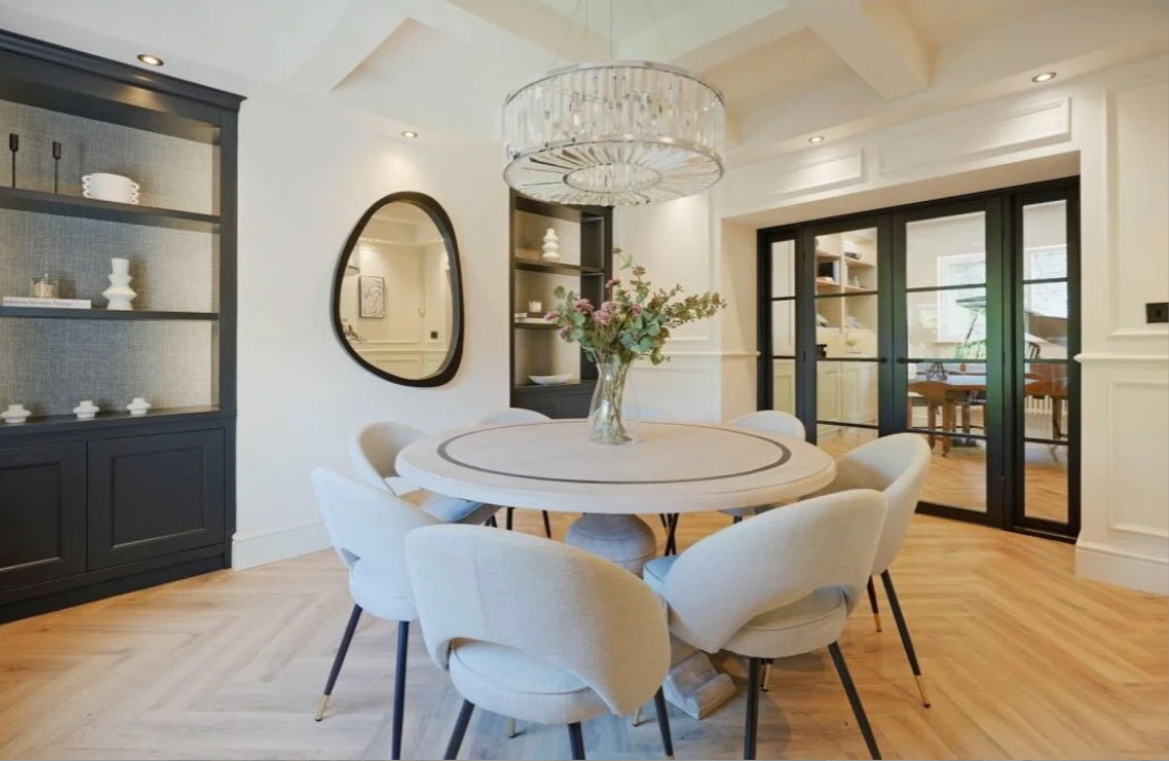 Modern dining room with a round table, six white upholstered chairs, a floral centerpiece, a large crystal chandelier, black sliding glass doors, built-in black shelves with decorative items, a wall mirror, and hardwood flooring.