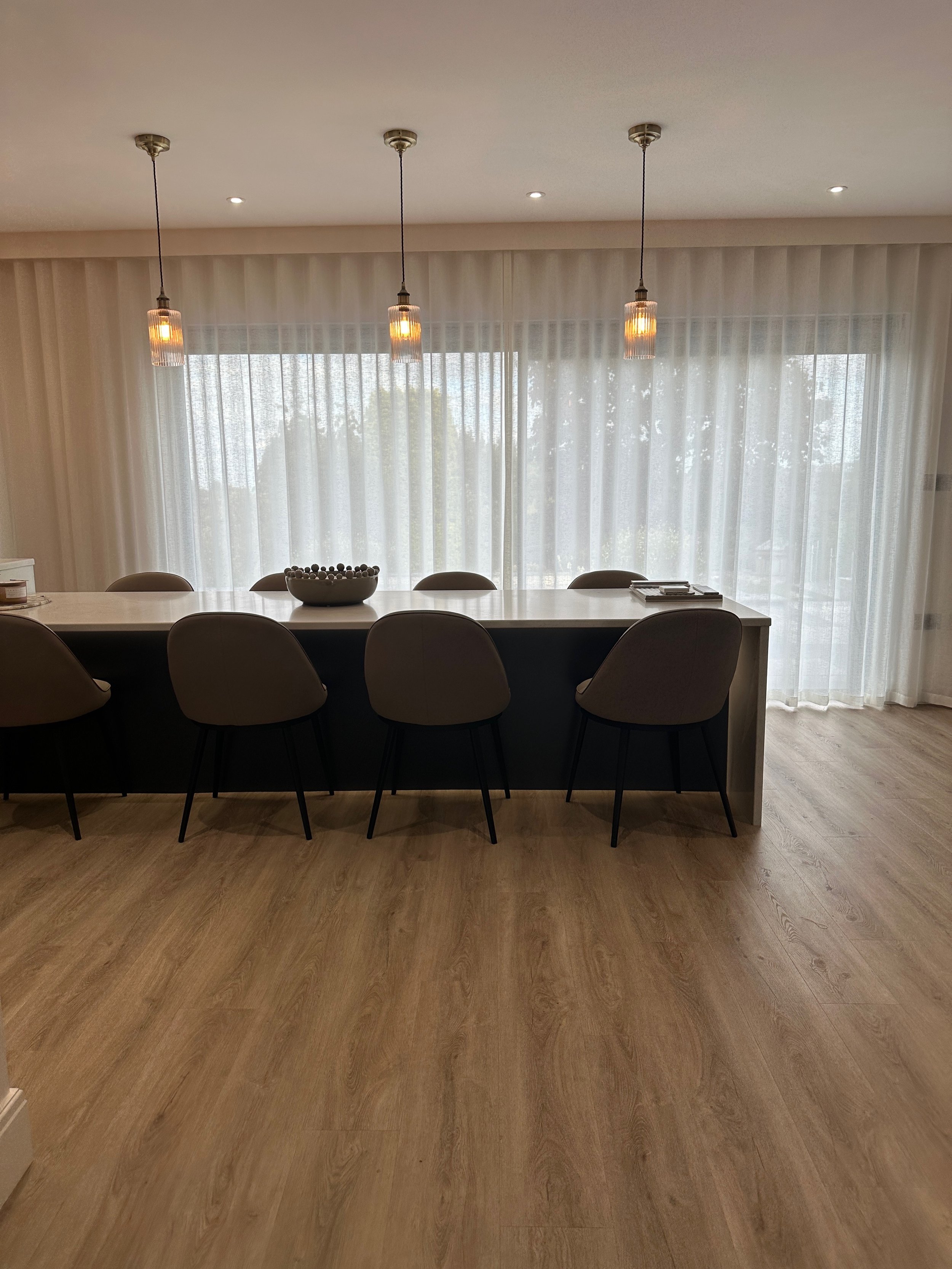 A dining area with a long table, five chairs, three pendant lights hanging from the ceiling, sheer curtains covering large windows, and a bowl on the table.