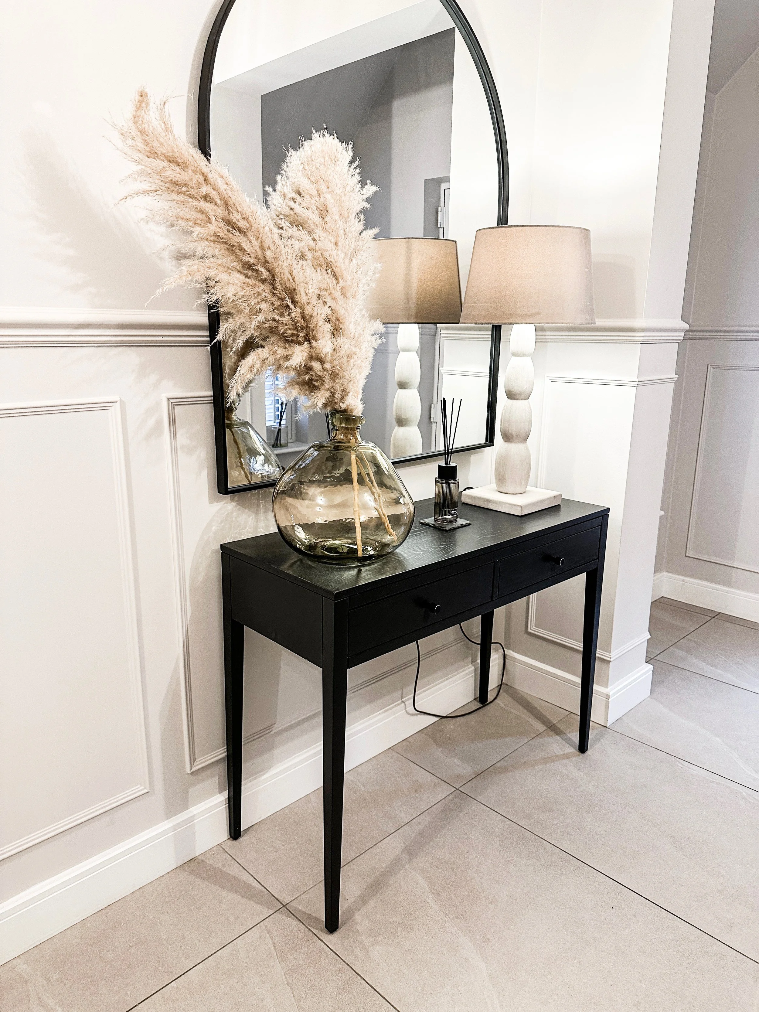 A black console table with a large glass vase holding pampas grass, two white table lamps with beige shades, a reed diffuser, and a mirror above reflects the scene in a modern, decorated entryway or living space.