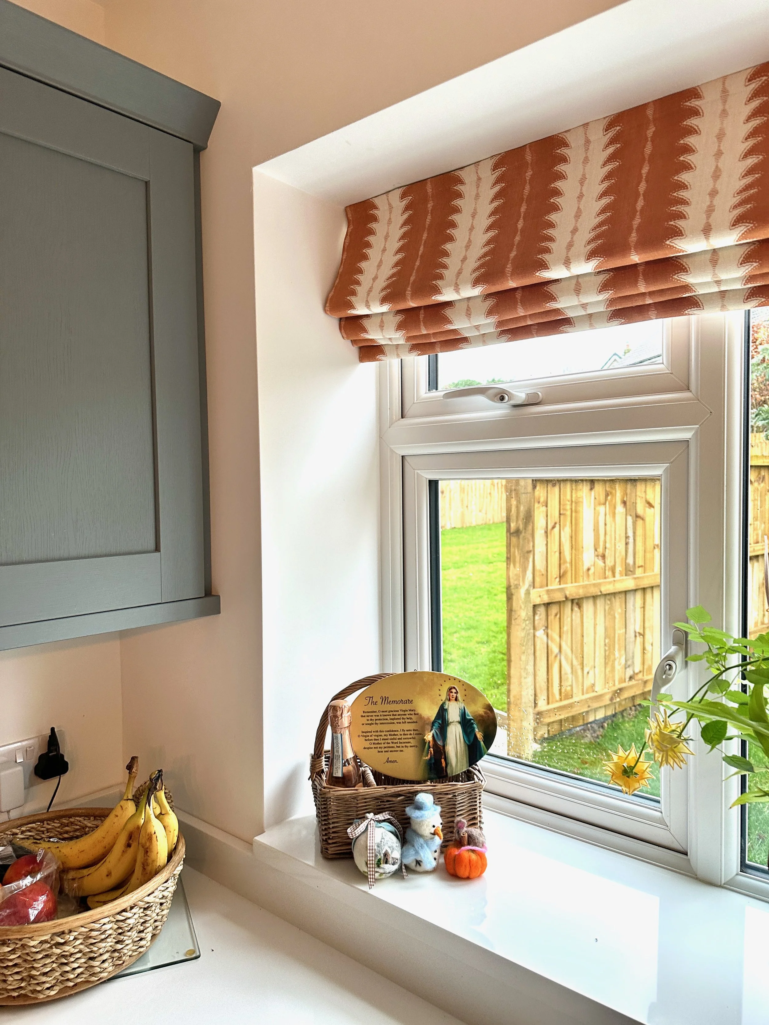 Kitchen windowsill with a basket of decorative items including a small pumpkin, plush snowman, and stuffed animals; some flowers in a pot; outside view of a wooden fence and grassy yard.