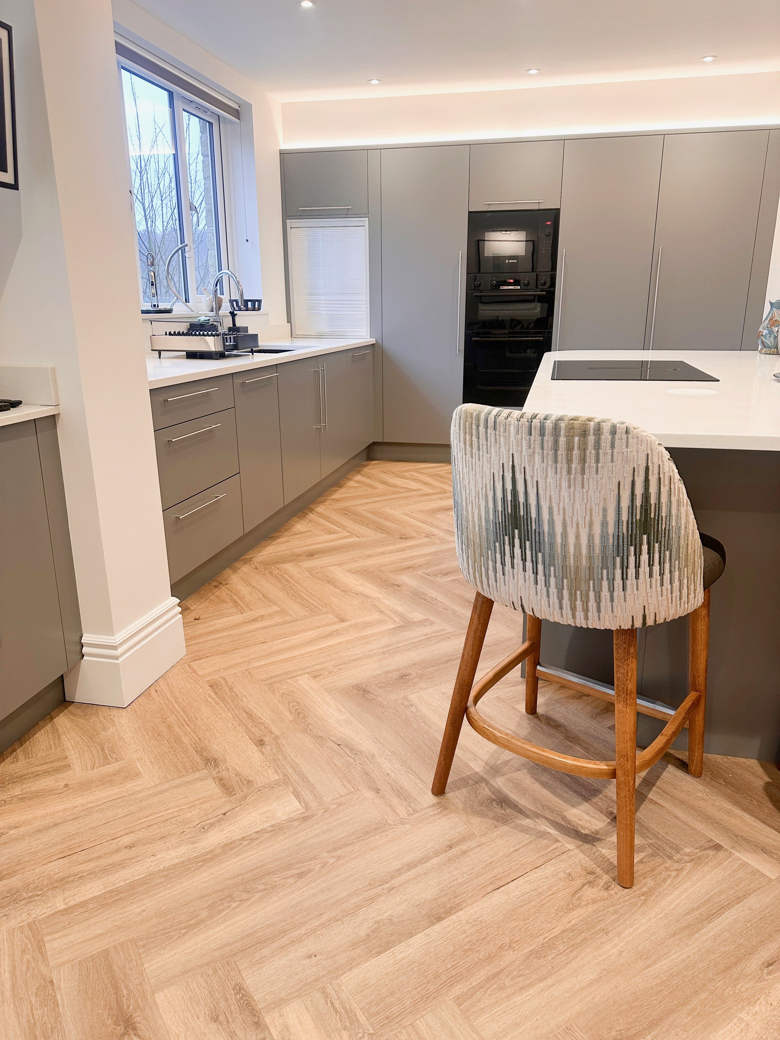 Modern kitchen with gray cabinets, white countertops, and wood-look flooring. Features a window with a view outside, an oven, and a bar stool with a patterned fabric seat at the island.