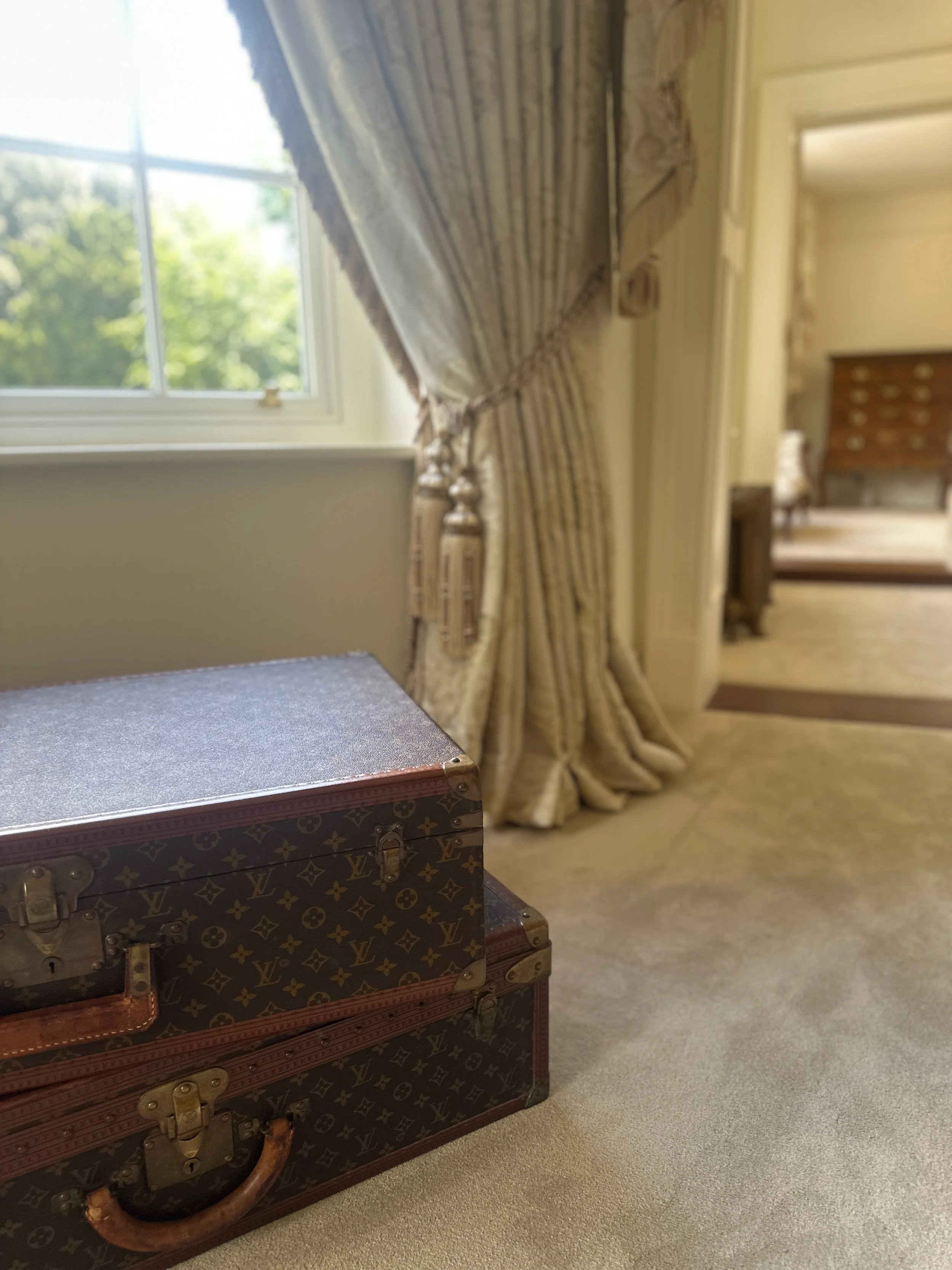 A vintage Louis Vuitton trunk sitting on a beige carpet in a room with large window and elegant curtains.