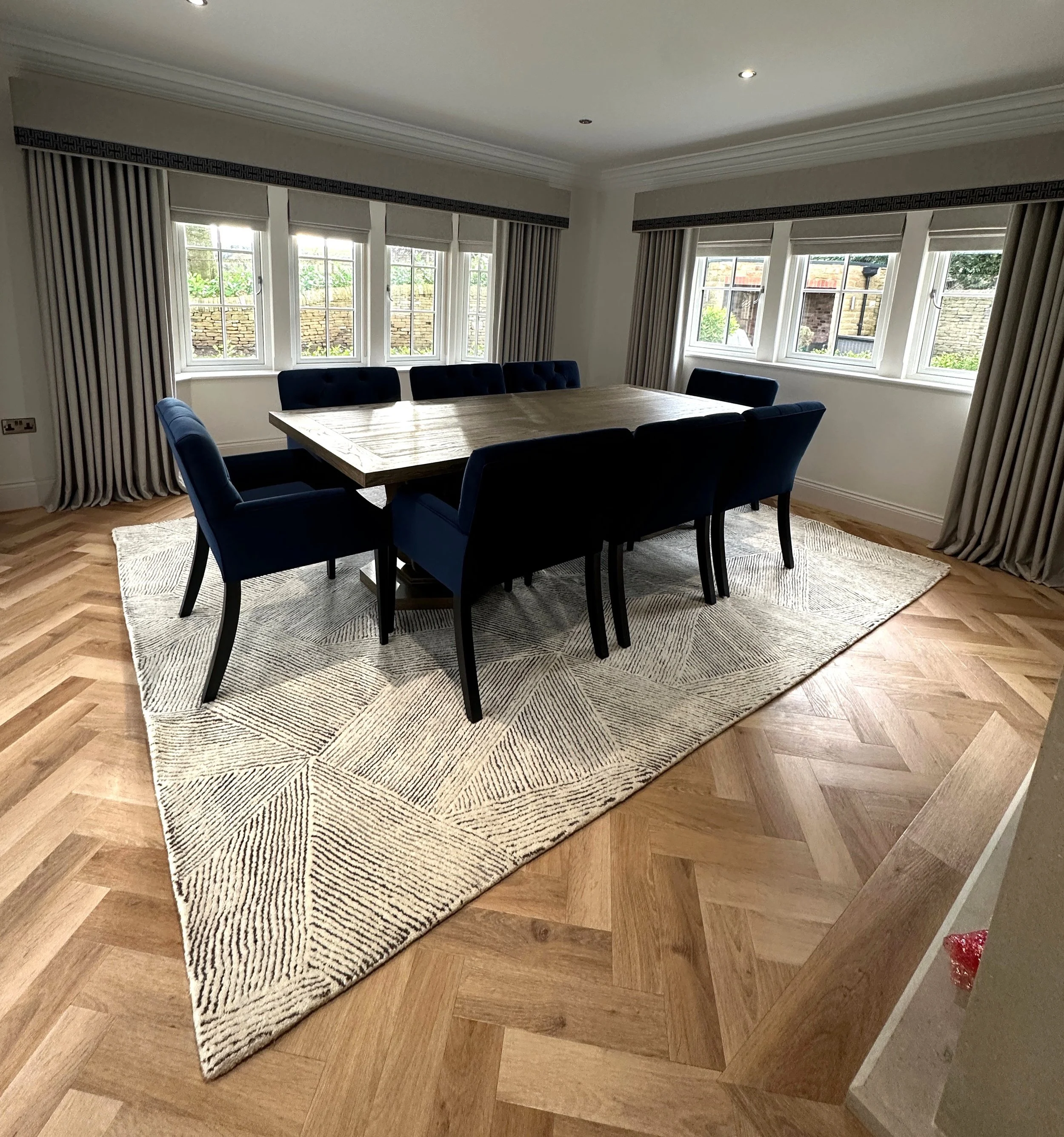 A spacious dining room with a large wooden table and enclosed navy blue chairs. The room has natural light coming through multiple windows, and beige curtains along with patterned area rug on wooden herringbone flooring.