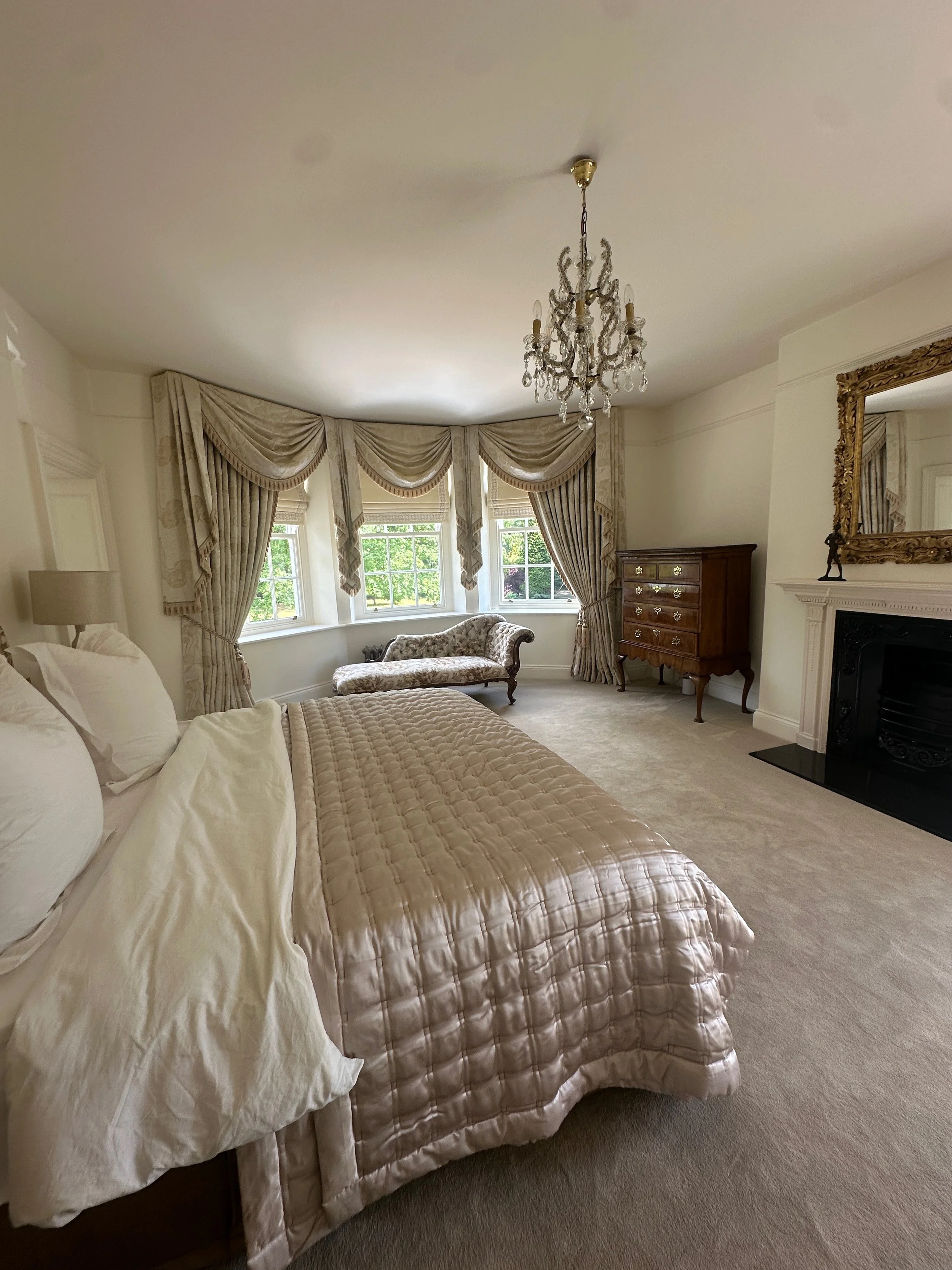 Elegant bedroom with a bed, vintage chaise lounge by windows, chandelier, ornate mirror, fireplace, and antique chest of drawers.