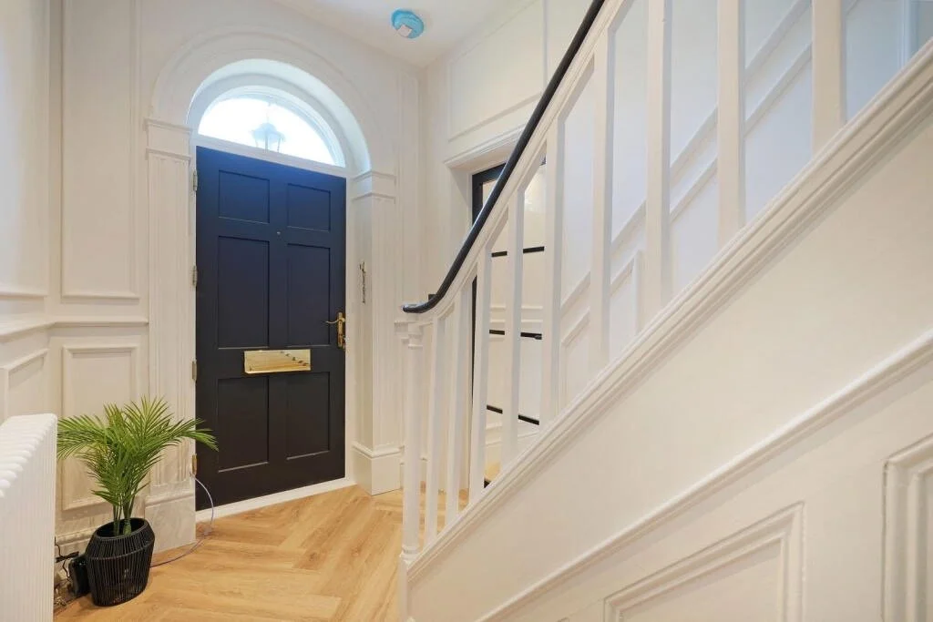 Entryway with a dark blue front door, a small potted green plant, white wainscoting, and a staircase with white spindles and black carpet treads.
