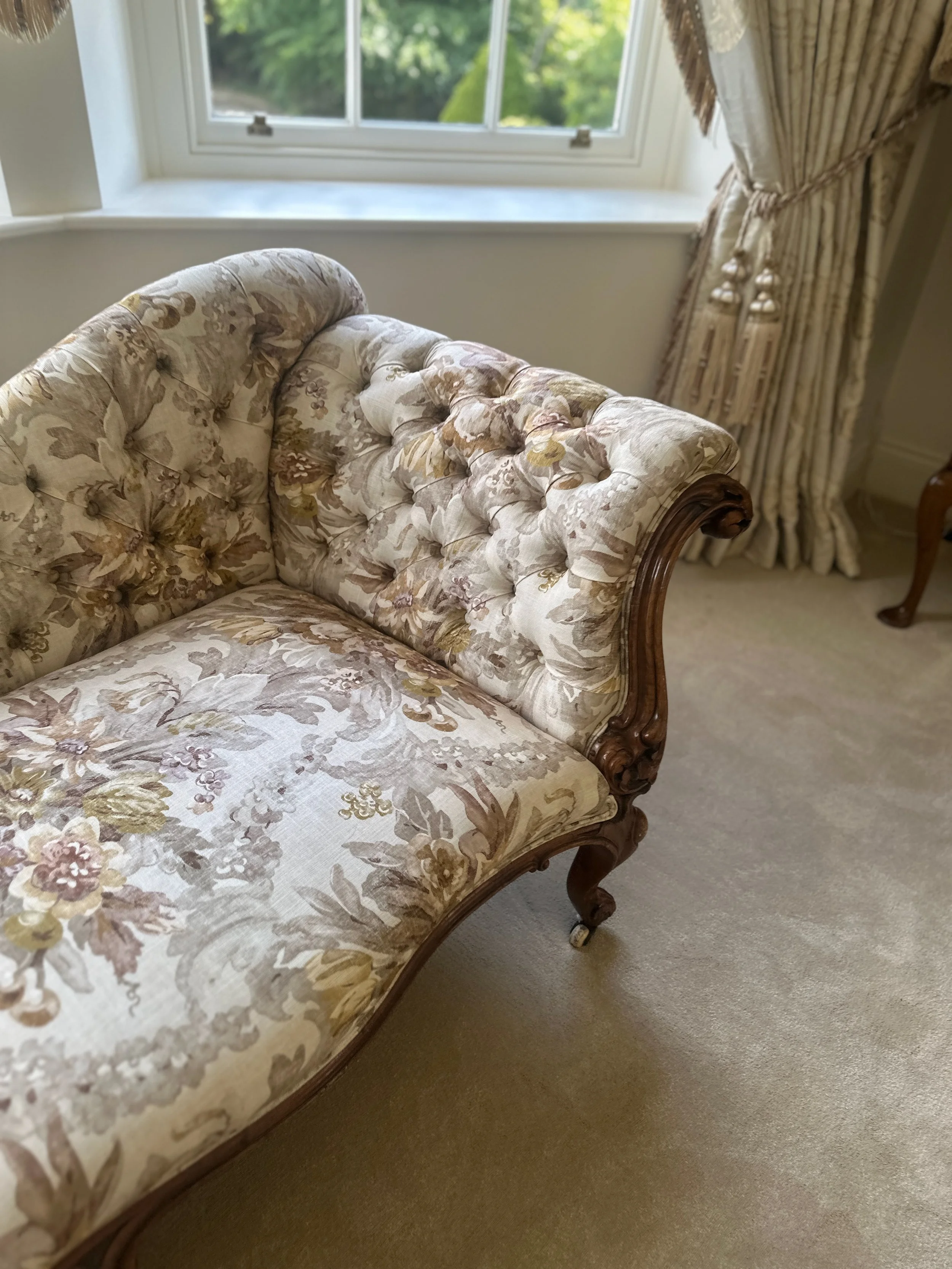 An ornate vintage-style sofa with floral upholstery and wooden carved accents, positioned in a room near a large window with draped curtains.