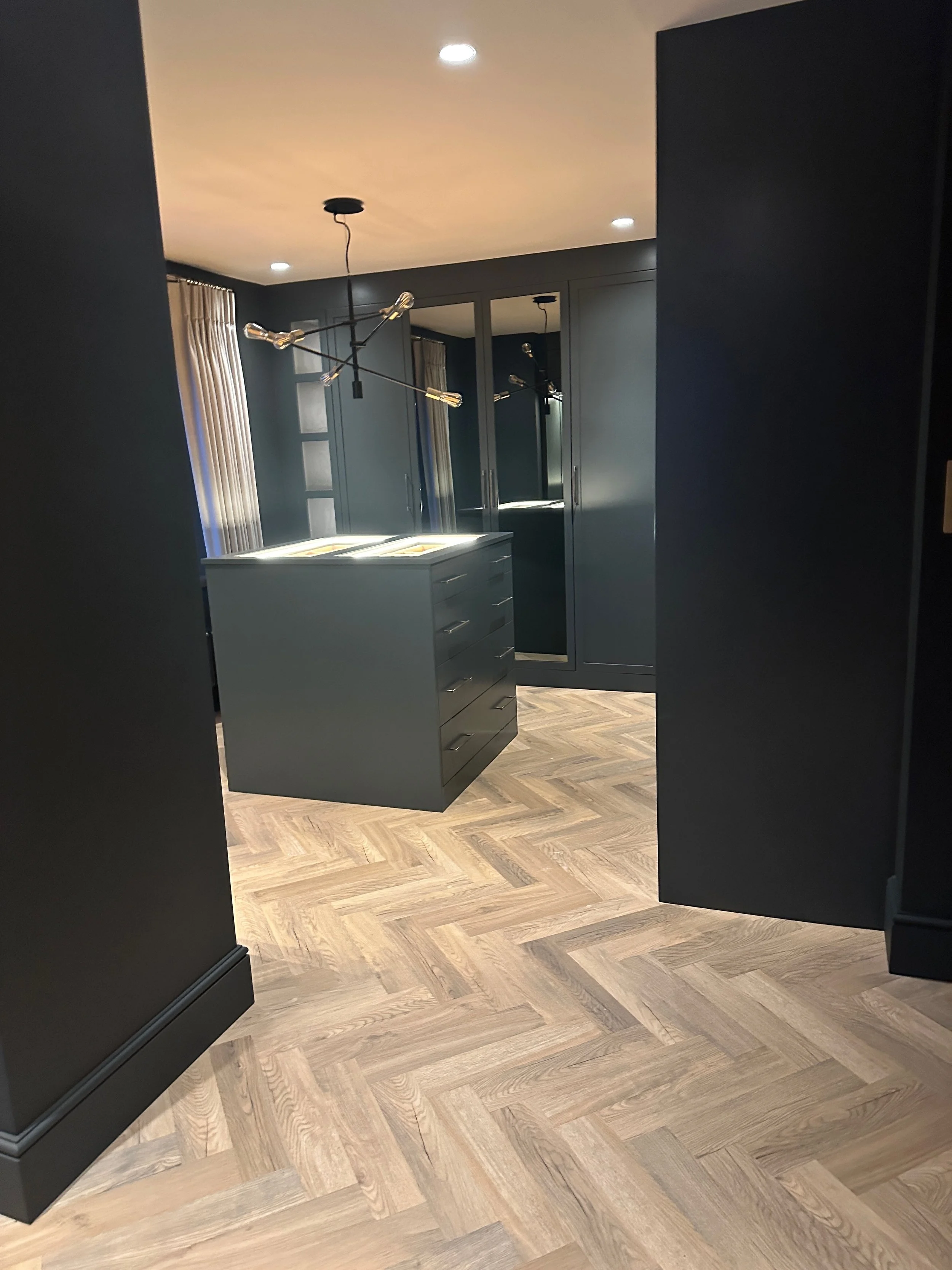 Modern interior space with dark walls, a matching dark gray island with drawers, a large closet with mirrored doors, and distressed wood herringbone flooring, illuminated by ceiling lights and a contemporary black chandelier.