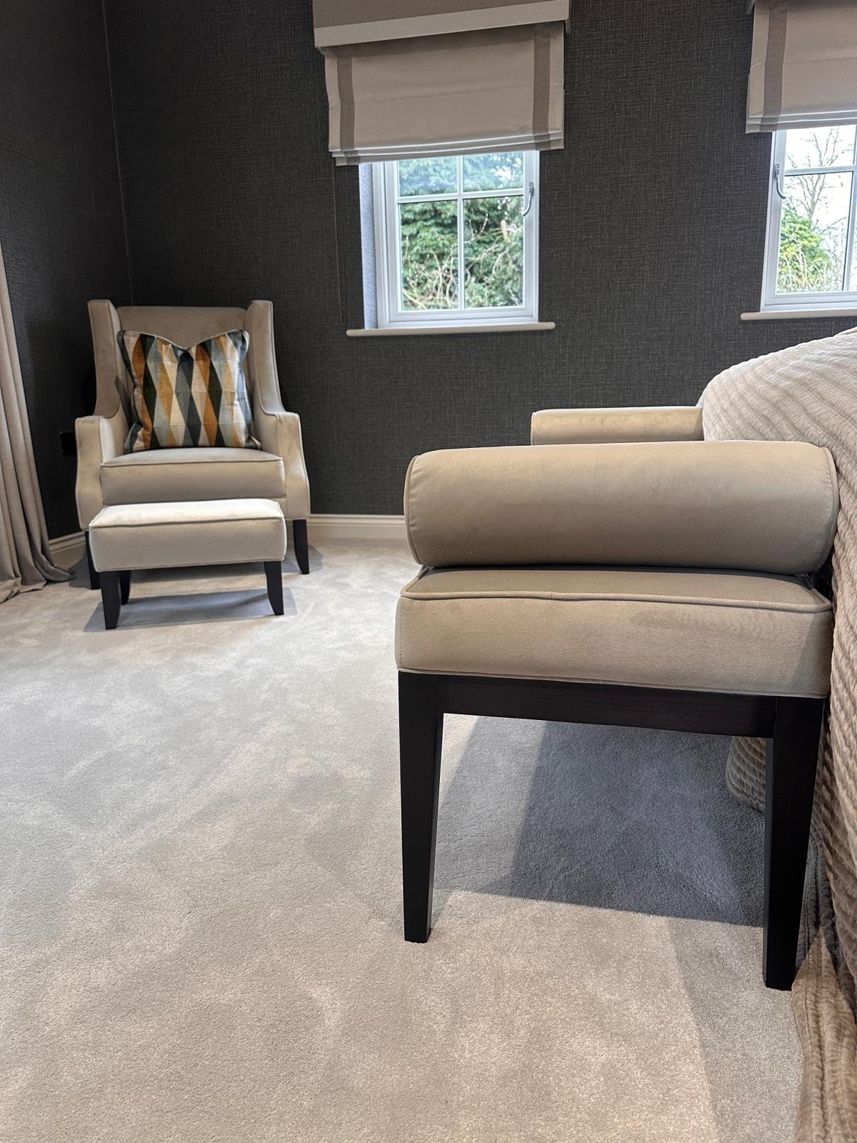 A cozy living room corner with gray walls, two windows with light-colored blinds, a white armchair with a decorative pillow, a matching ottoman, and a beige sofa with a light-colored blanket.