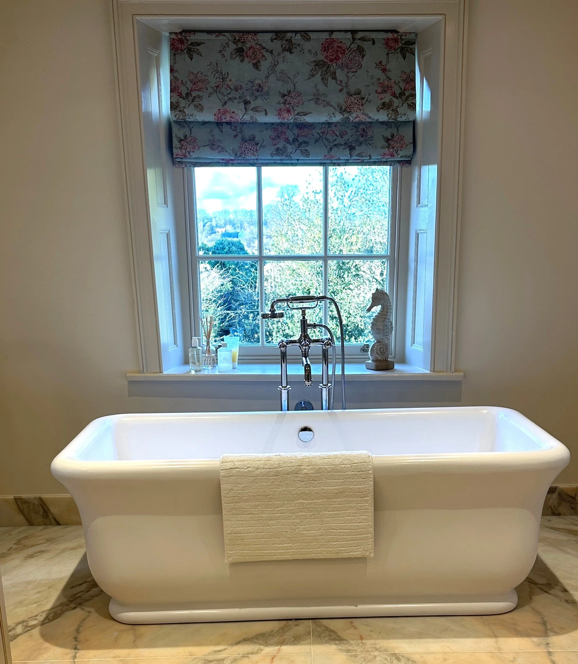 A white freestanding bathtub with a towel draped over its edge, positioned in front of a window with a floral valance. The window shows a view of trees and a partly cloudy sky. On the window sill, there are decorative items including a seahorse sculp