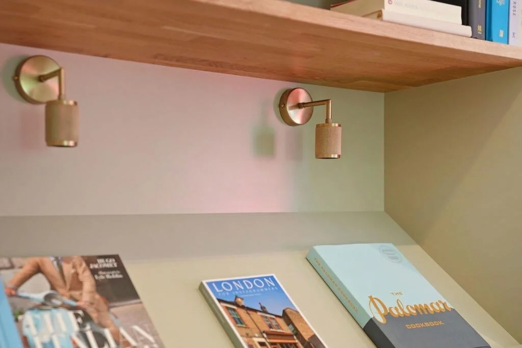 Close-up of wall-mounted adjustable reading lights above a desk with travel books, including 'London' and 'The Palomar Cookbook'.