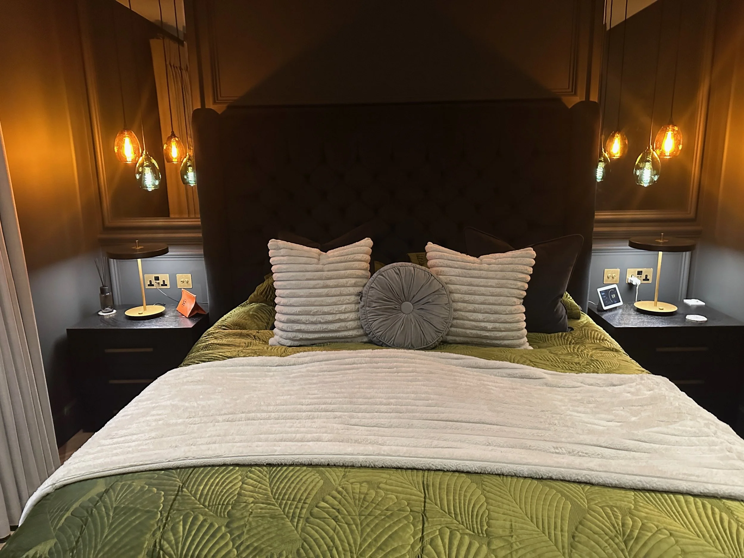 A neatly made bed with green quilt and white throw, six decorative pillows, two bedside tables with lamps and electronic devices, pendant lights hanging on dark wood paneled walls.