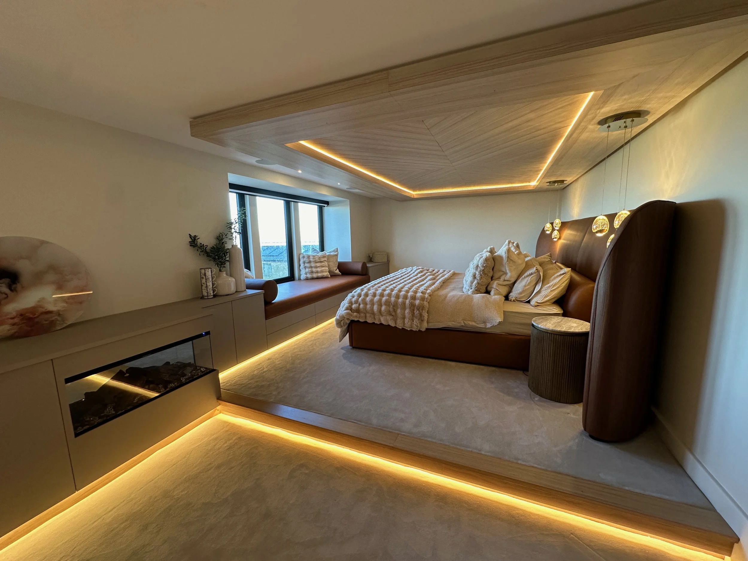 Modern bedroom with a large brown bed with pillows and blankets, built-in window seat with cushions by a window, contemporary ceiling design with LED strip lighting, and warm yellow accent lighting along the floor and furniture.