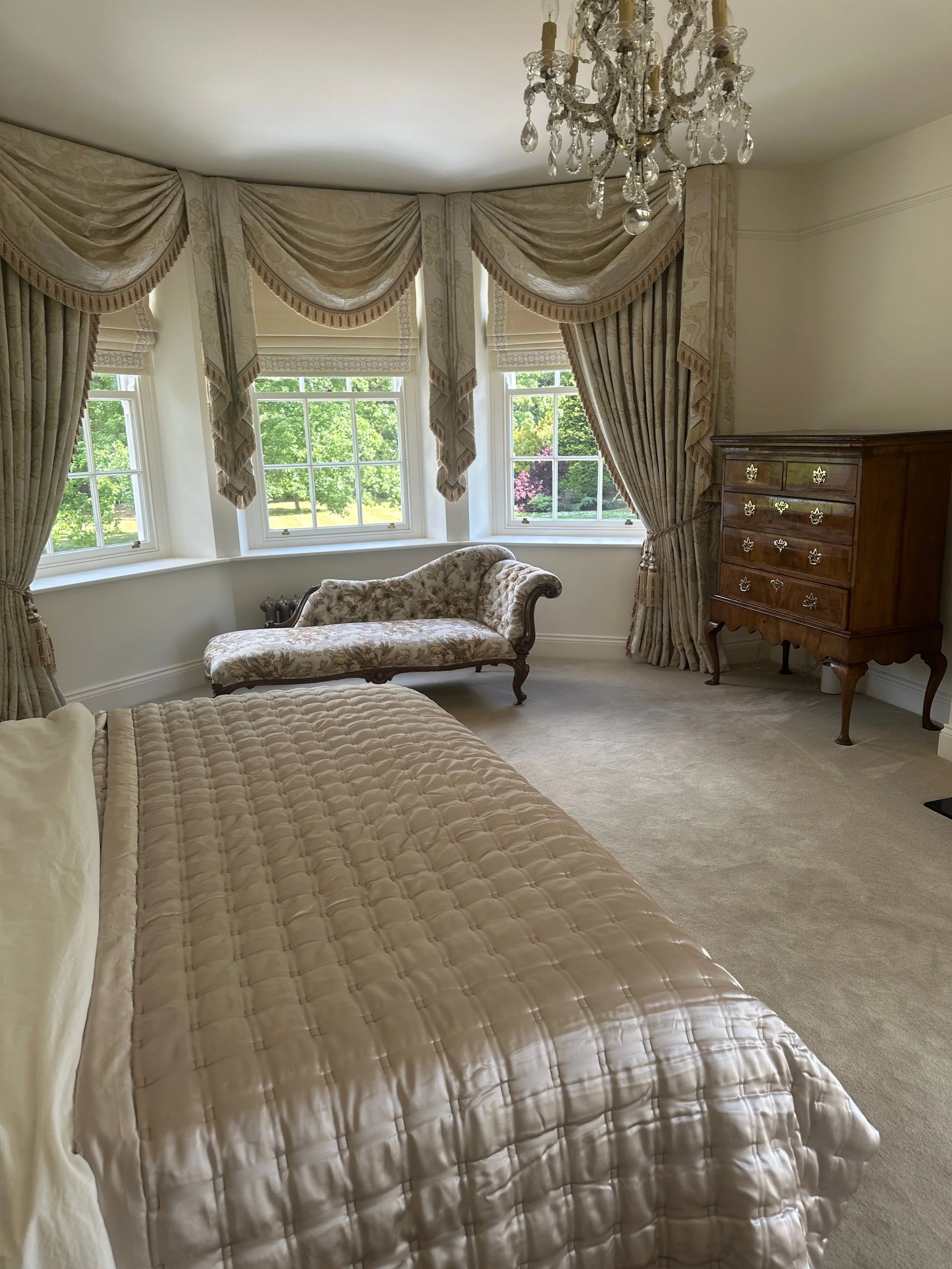 Elegant bedroom with a large bed covered in a shiny beige quilt, vintage-style chaise lounge with floral upholstery near three windows with elaborate gold and beige drapes, a crystal chandelier hanging from the ceiling, and a wooden dresser with gold