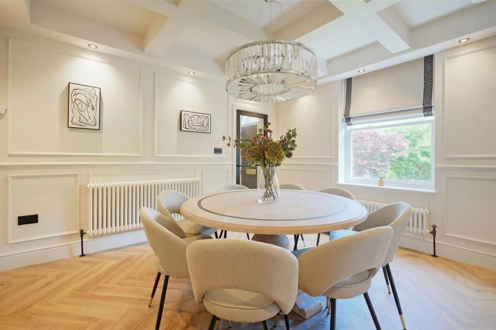 A bright dining room with a round table and eight beige chairs, a flower vase centerpiece, a large window, a chandelier, white paneled walls, and wooden flooring.