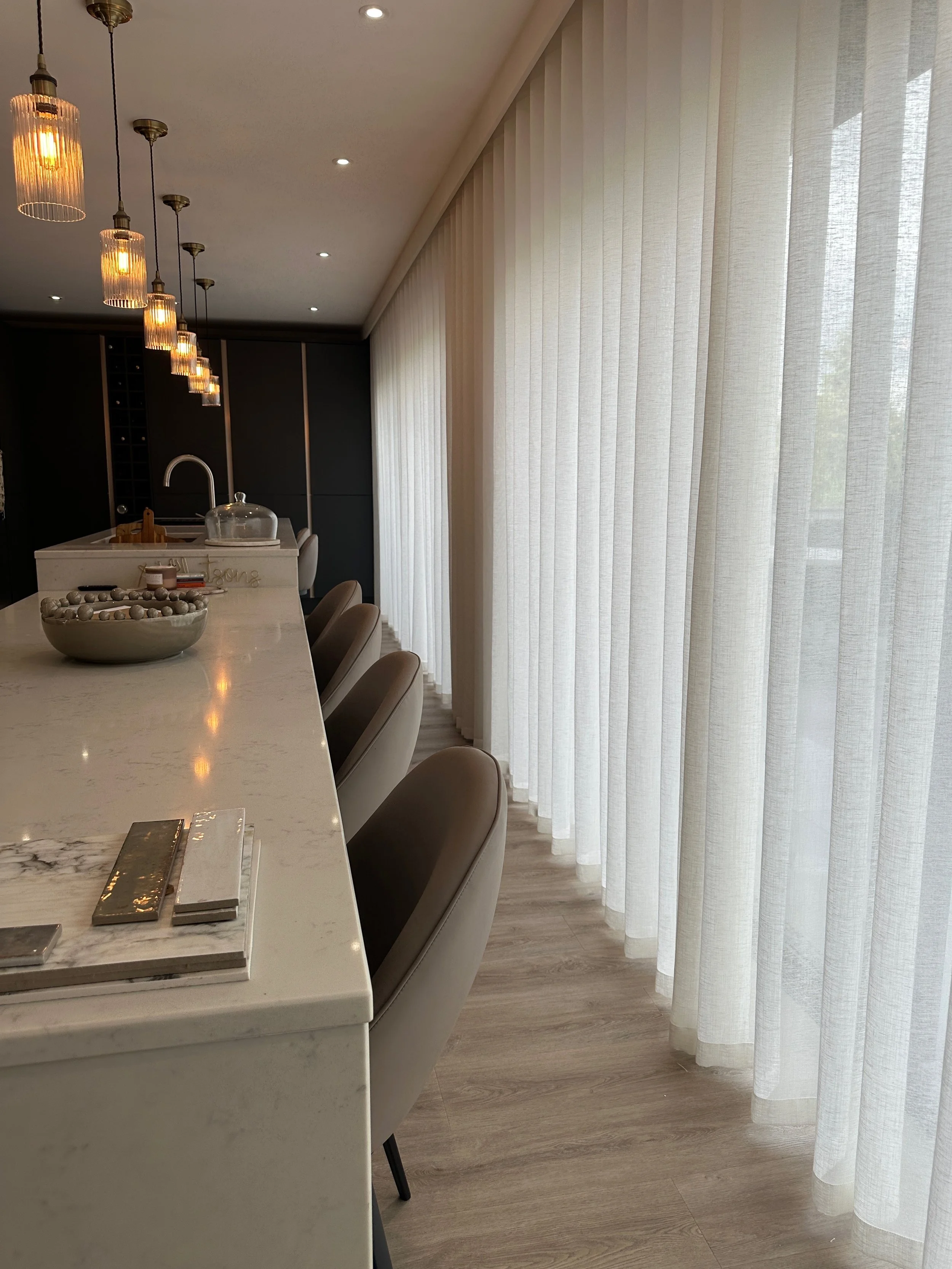 Modern kitchen with a white marble island, beige chairs, large windows with white curtains, and a black accent wall with pendant lighting.