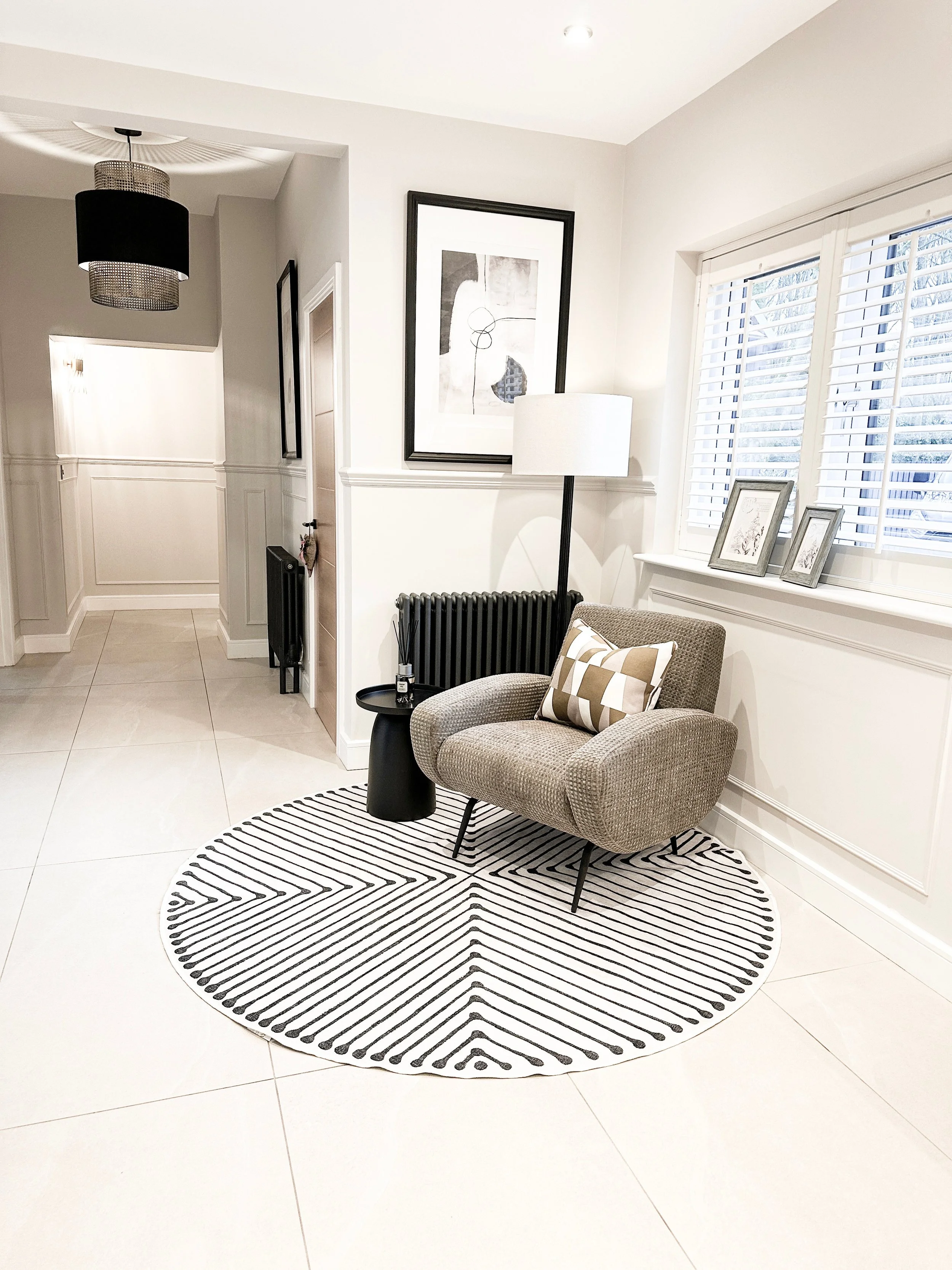 Living room corner with a cozy armchair, black radiator, floor lamp, framed artwork, and window with white shutters, all on a black and white patterned round rug.