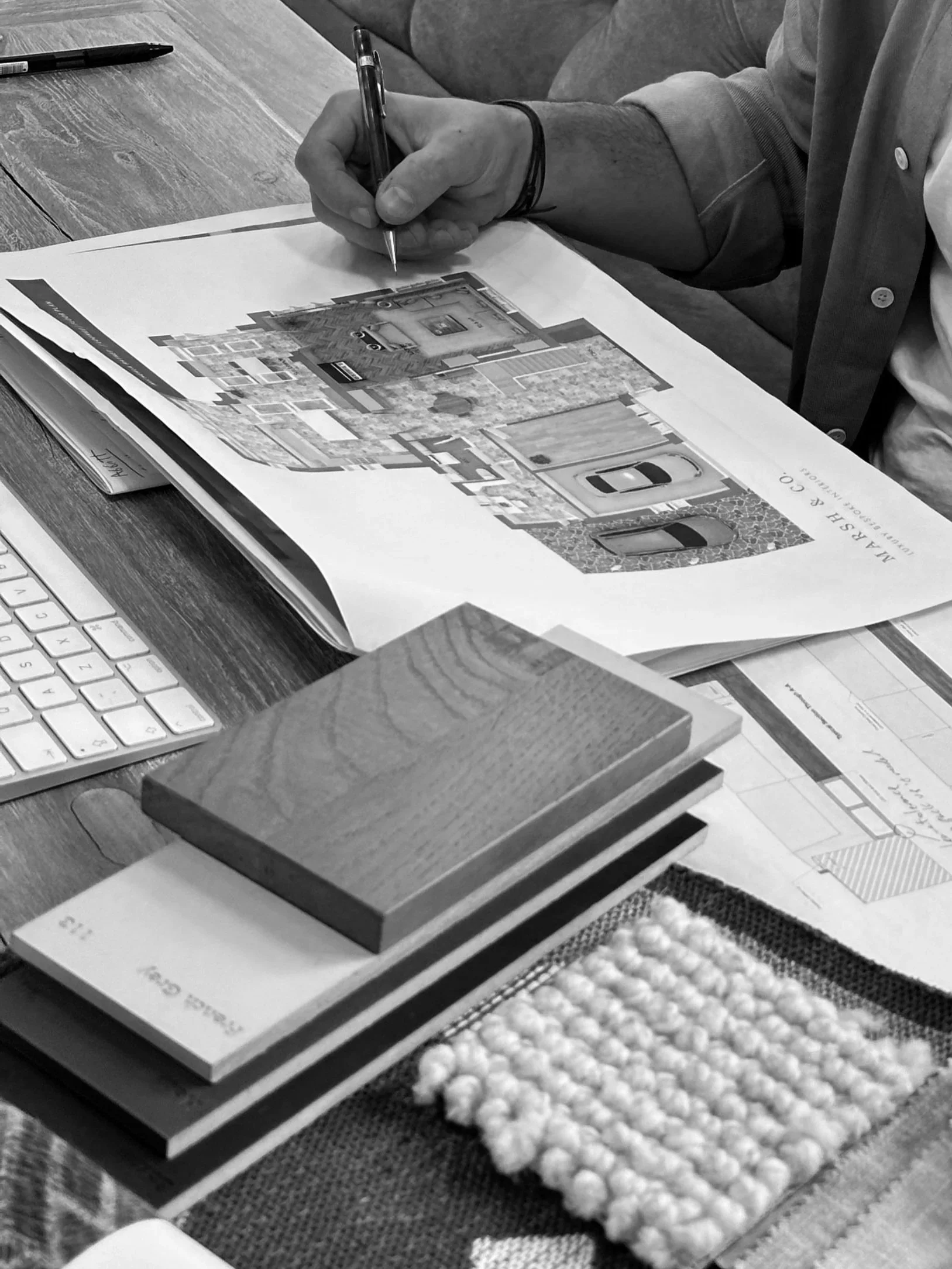 Person reviewing a design or floor plan at a wooden table, with color swatches, fabric samples, and a keyboard nearby.