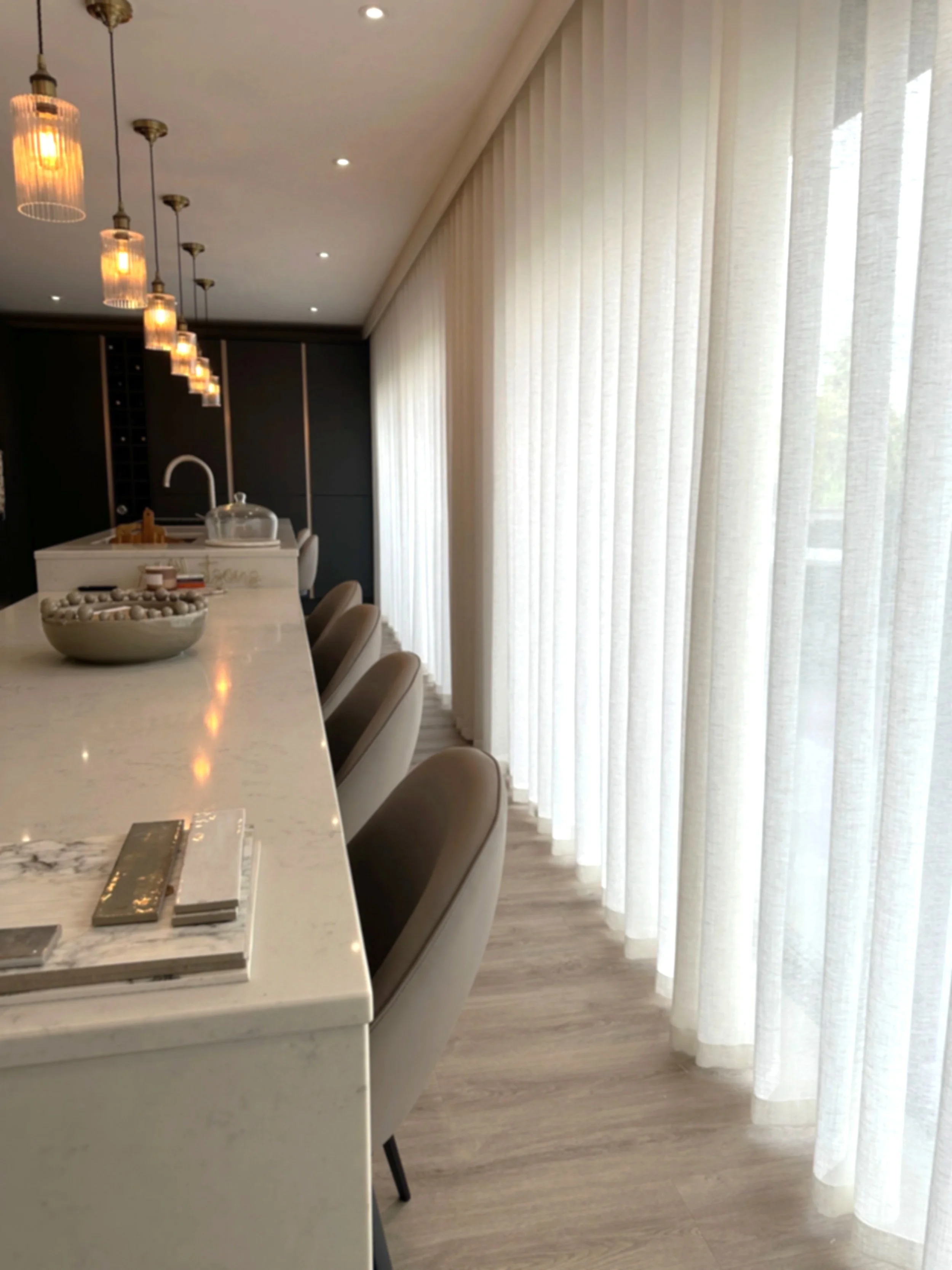 Modern kitchen with a marble island, beige chairs, pendant lighting, and sheer curtains covering large windows.