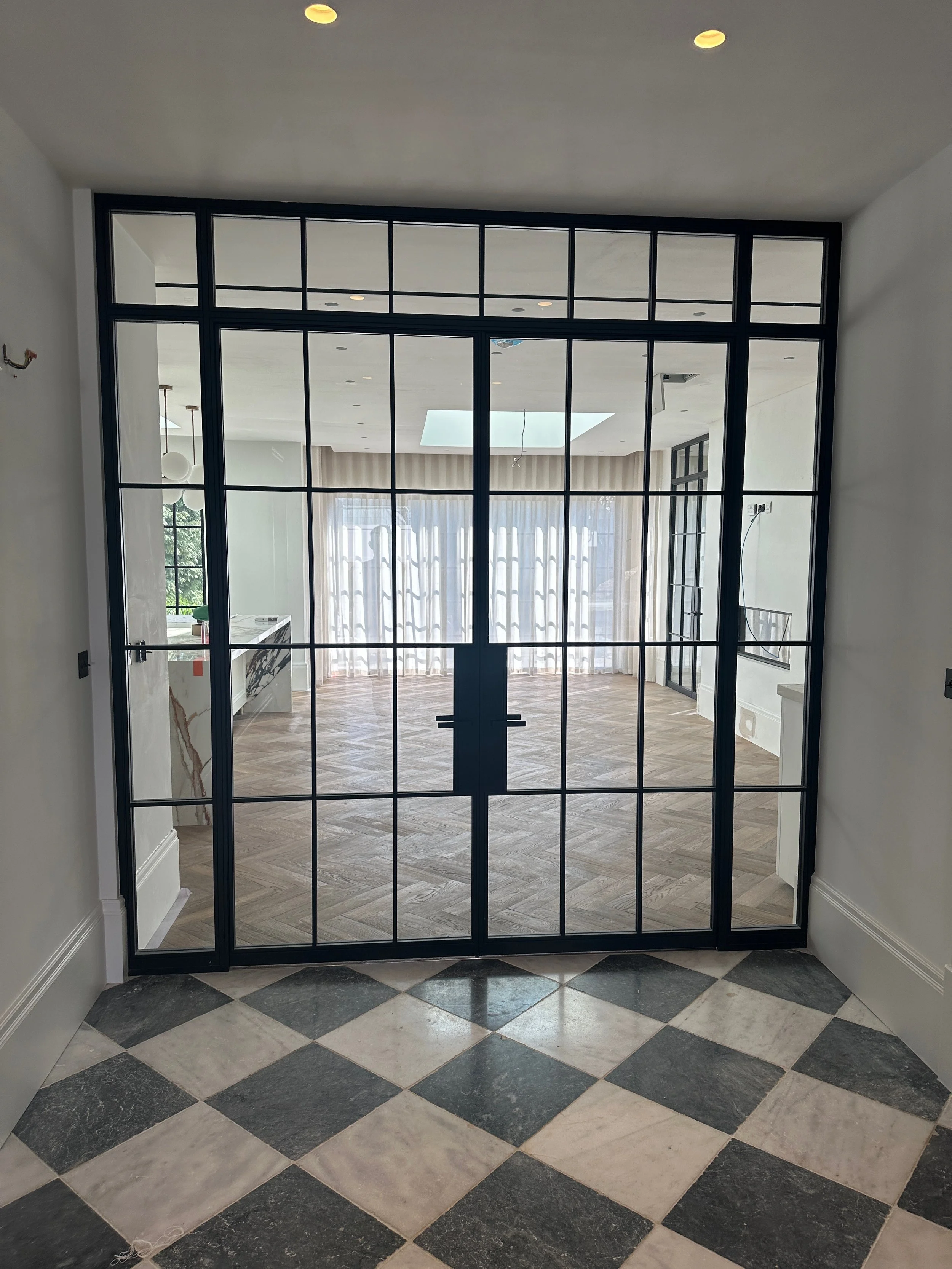 Interior view through a large glass and metal door, showing a spacious room with wooden flooring, white walls, and large windows with curtains.