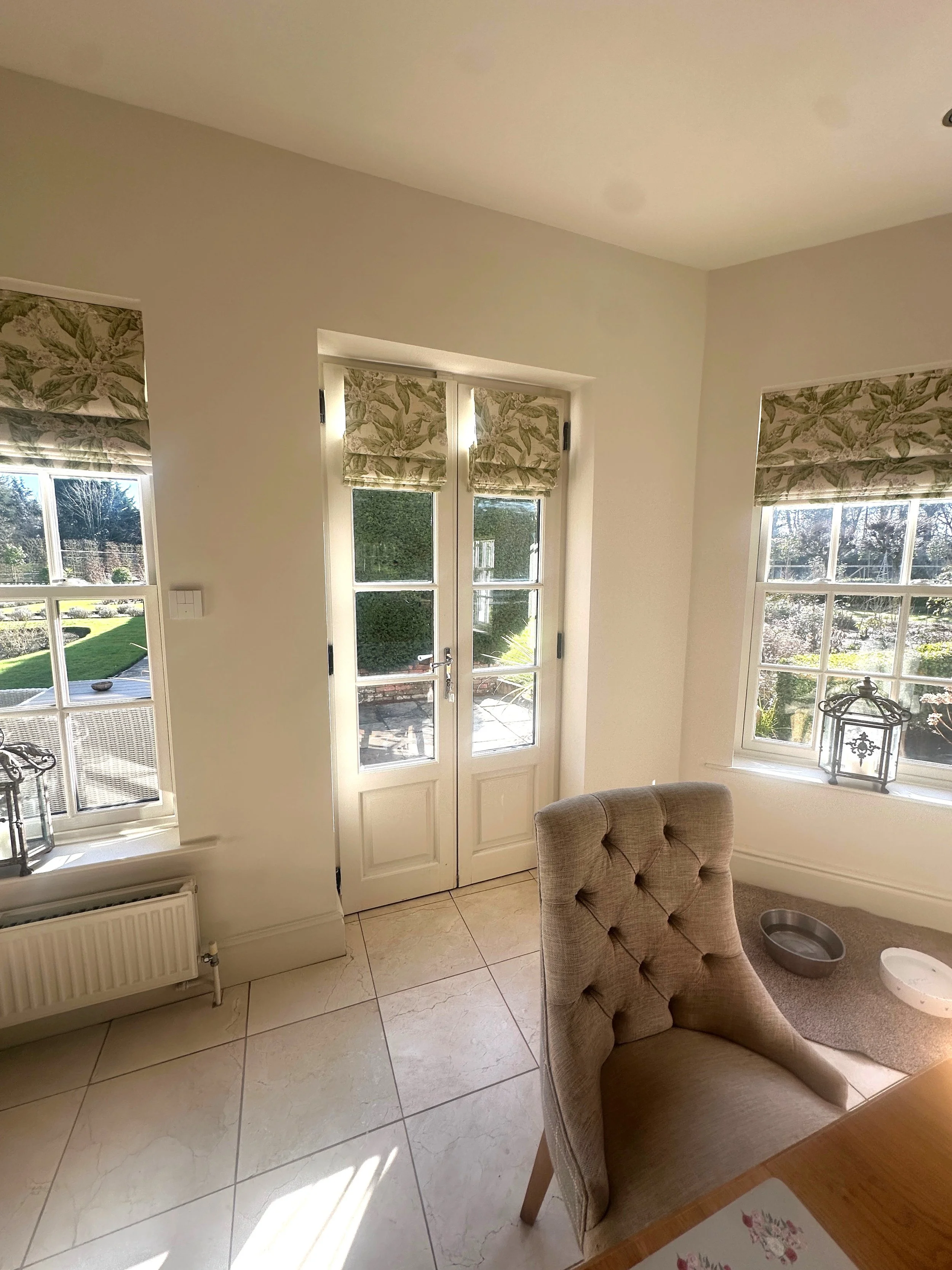 A bright dining area with double glass doors leading outside, surrounded by windows with patterned roman blinds, a beige upholstered chair, and a small pet water dish on a mat on the floor.