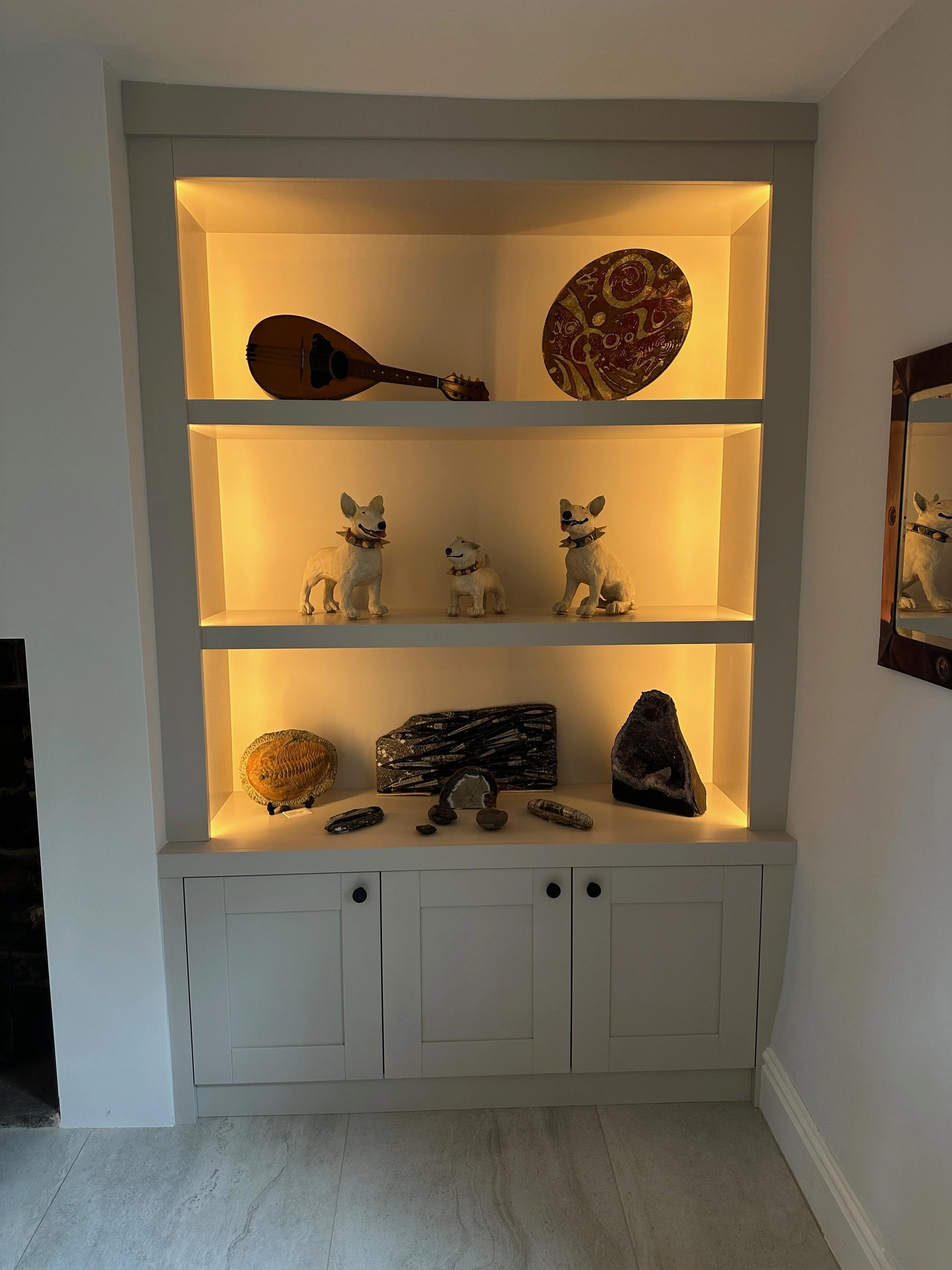 Decorative display shelf with musical instrument, paintings, dog figurines, and stones, illuminated with warm yellow lighting.