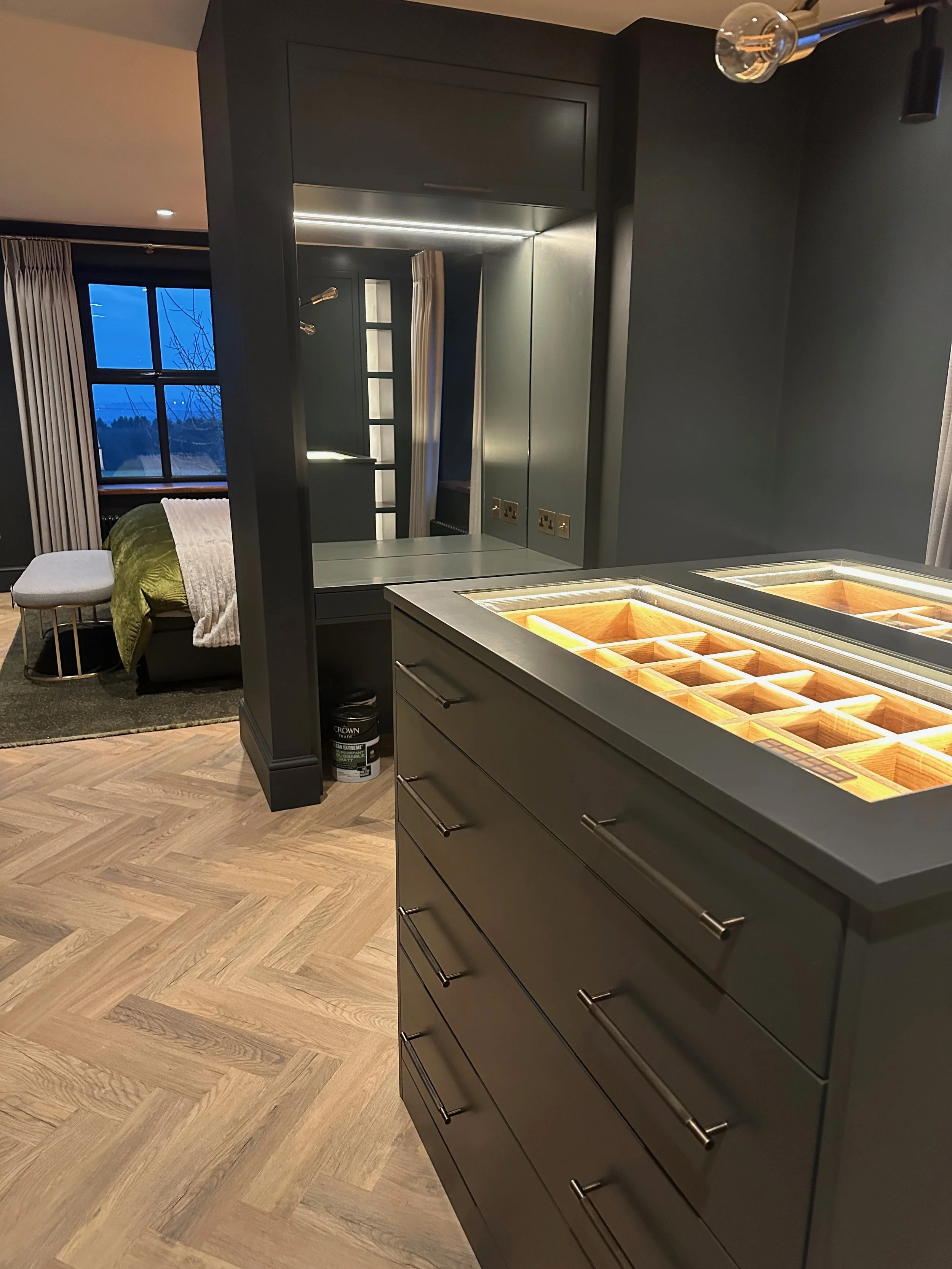 Modern open-plan living area with a bedroom in the background, featuring a window with curtains, a bed with green and white bedding, and a small grey ottoman. The foreground shows a dark grey cabinet with a lit glass top containing wooden compartment