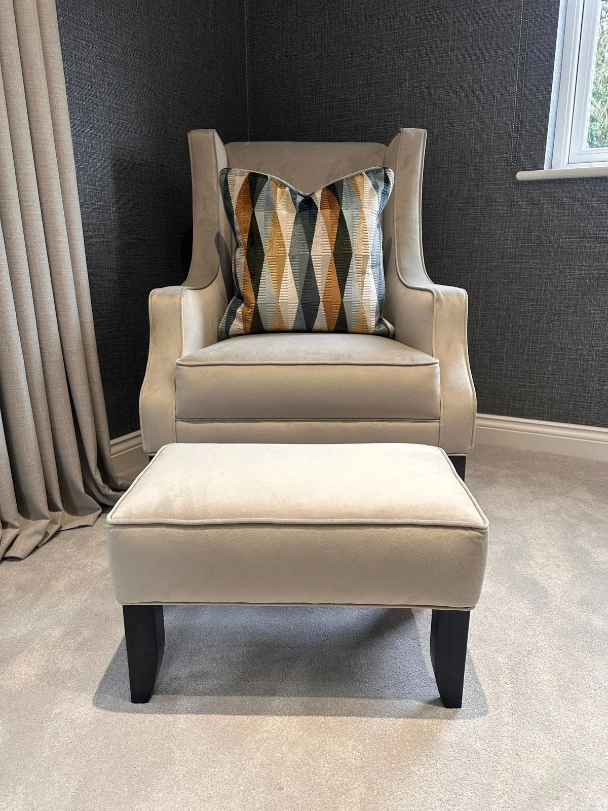 A beige upholstered armchair with a decorative pillow featuring a geometric pattern in shades of black, tan, gray, and beige, situated next to a window with beige curtains, against a dark textured wall and a light-colored carpet.