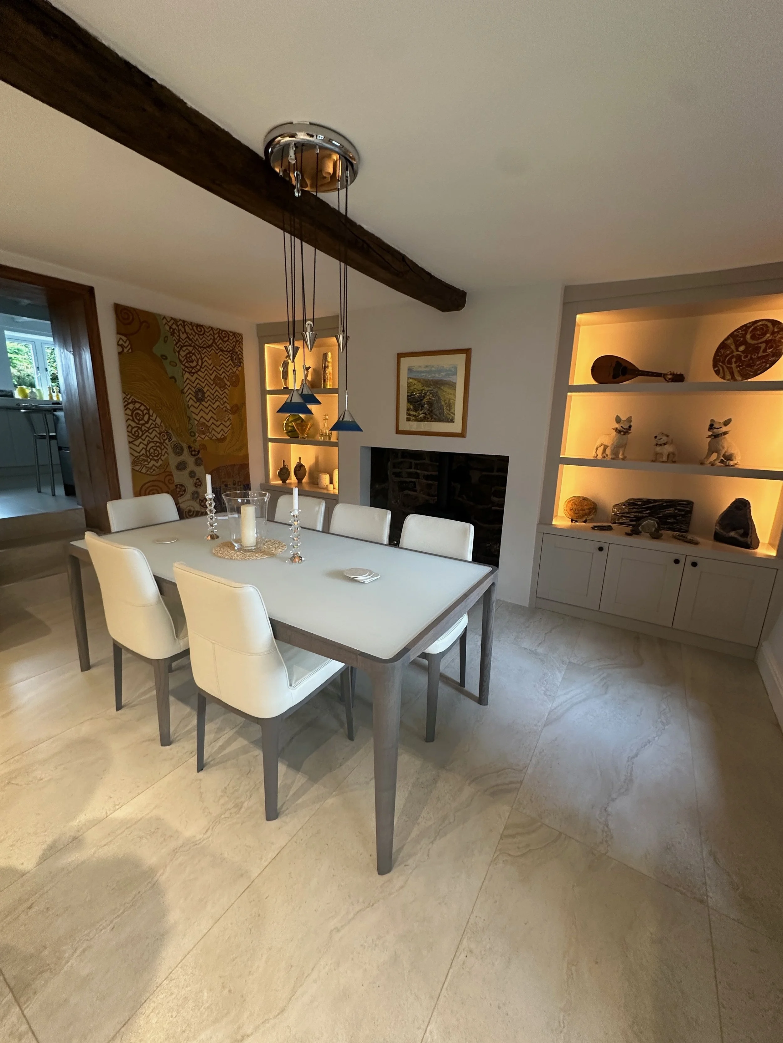 Dining room with white table and six white chairs, artwork on the wall, built-in shelves with decorative items, wooden ceiling beam, and tiled floor.