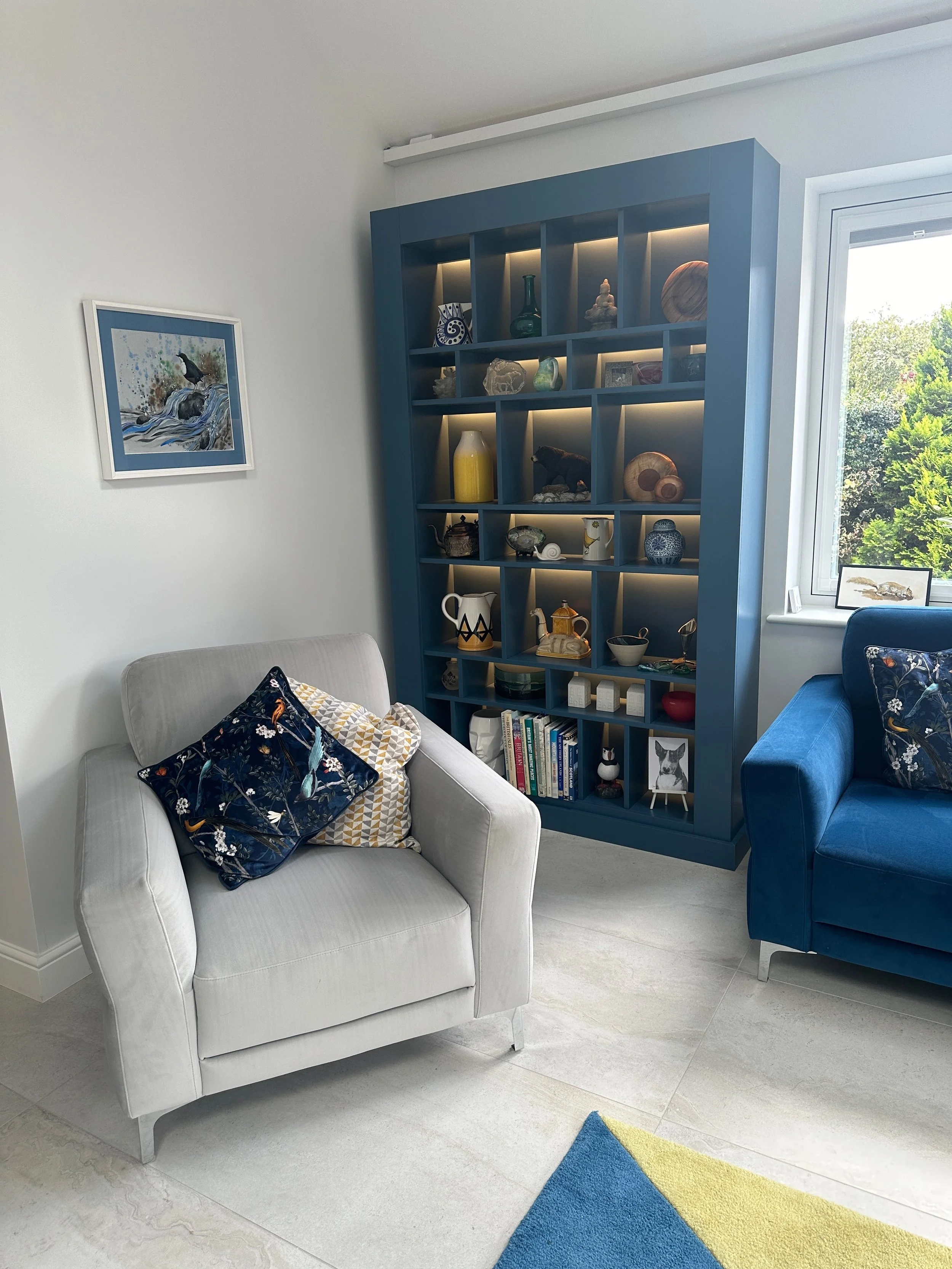 Living room with light gray armchair, navy blue bookshelf displaying decorative items, blue sofa with patterned throw pillow, window with view of green trees, and a colorful rug.