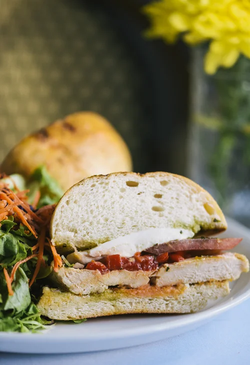 A photo of a sandwich cross-section from Farm Country Kitchen. Featuring grilled chicken, red pepper, tomato, pesto, on a fluffy bread; with a side of mixed greens and shredded carrot.