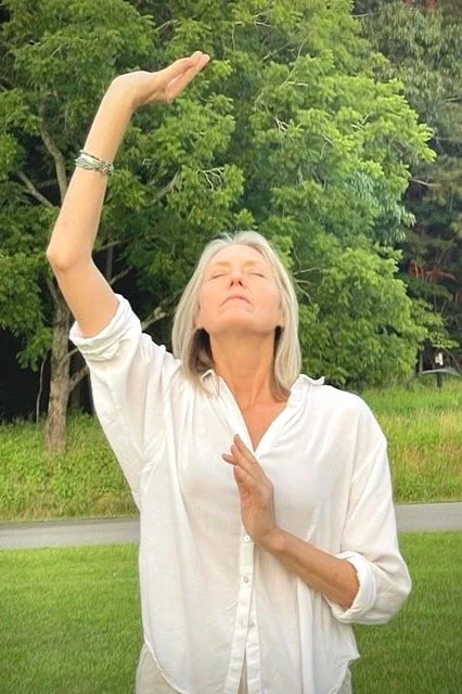 A woman with gray hair in a white shirt standing outdoors with trees in the background..