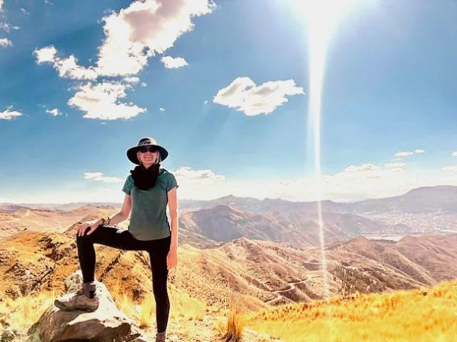 A woman standing on a rock in a mountainous landscape with clear blue sky and scattered clouds, wearing sunglasses, a wide-brimmed hat, a teal shirt, and black pants.