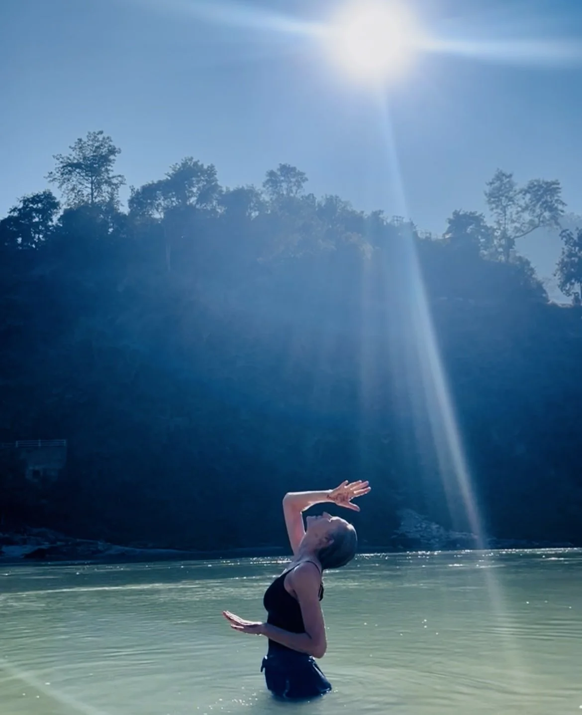 Woman standing in a river with her head tilted back, arms raised above head, during bright sunny day with trees on a hillside in the background.