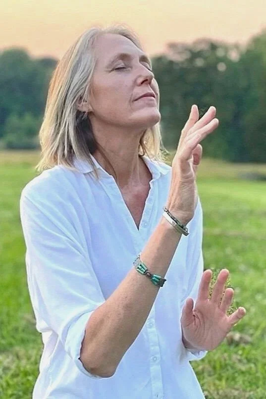An older woman with gray hair is outdoors, wearing a white shirt, with eyes closed at sunset.
