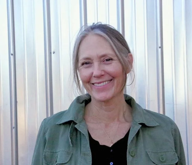 A woman with gray hair smiling, wearing a green jacket over a black top, standing in front of a metallic wall.