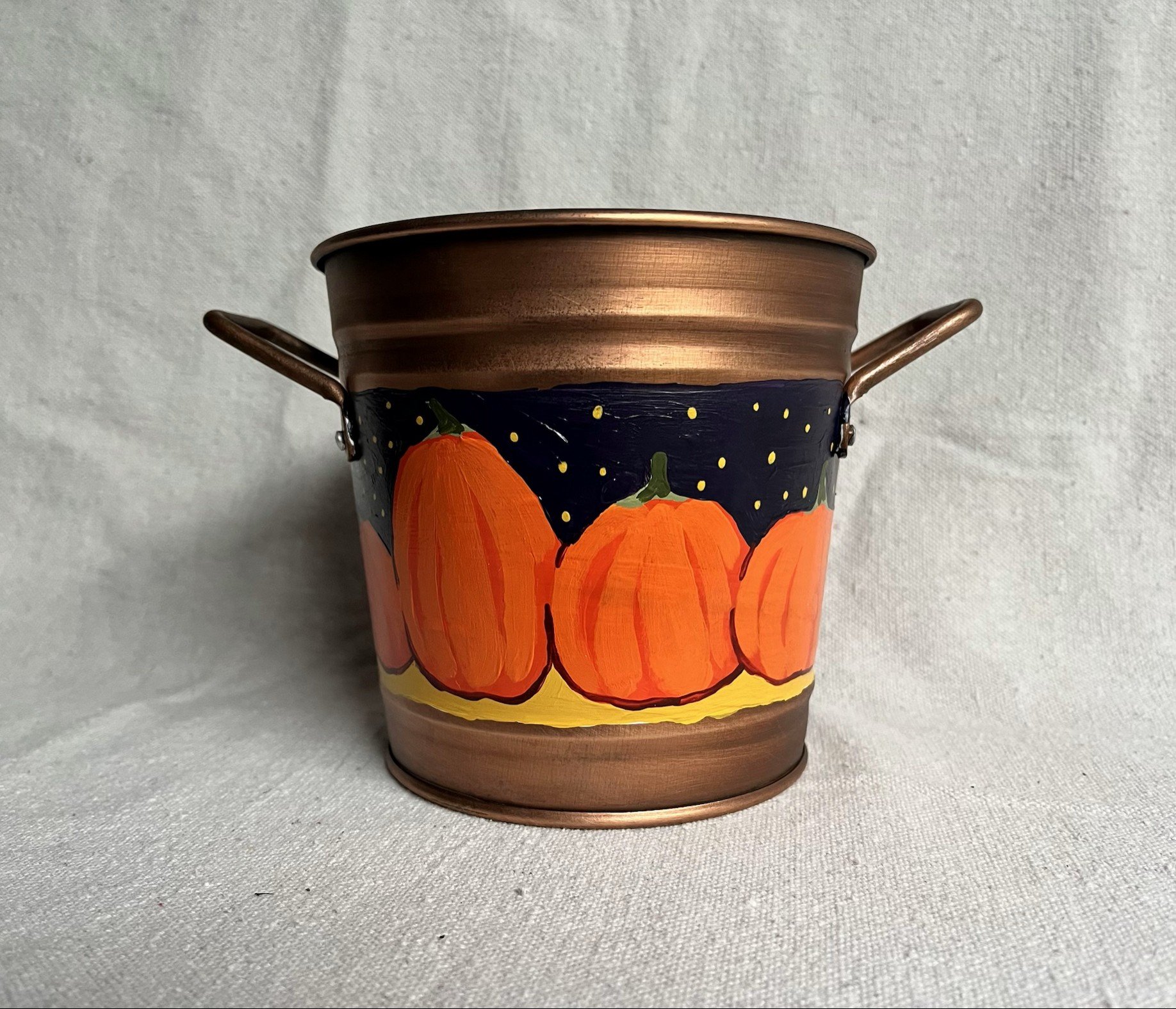 A hand painted pumpkin patch against a starry night sky, painted on a copper color tin bucket.