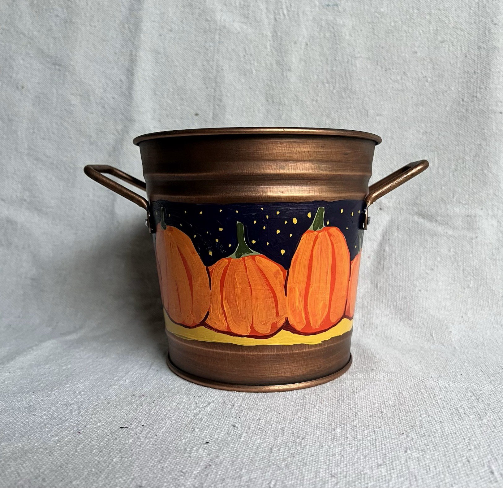 A hand painted pumpkin patch against a starry night sky, painted on a copper color tin bucket.