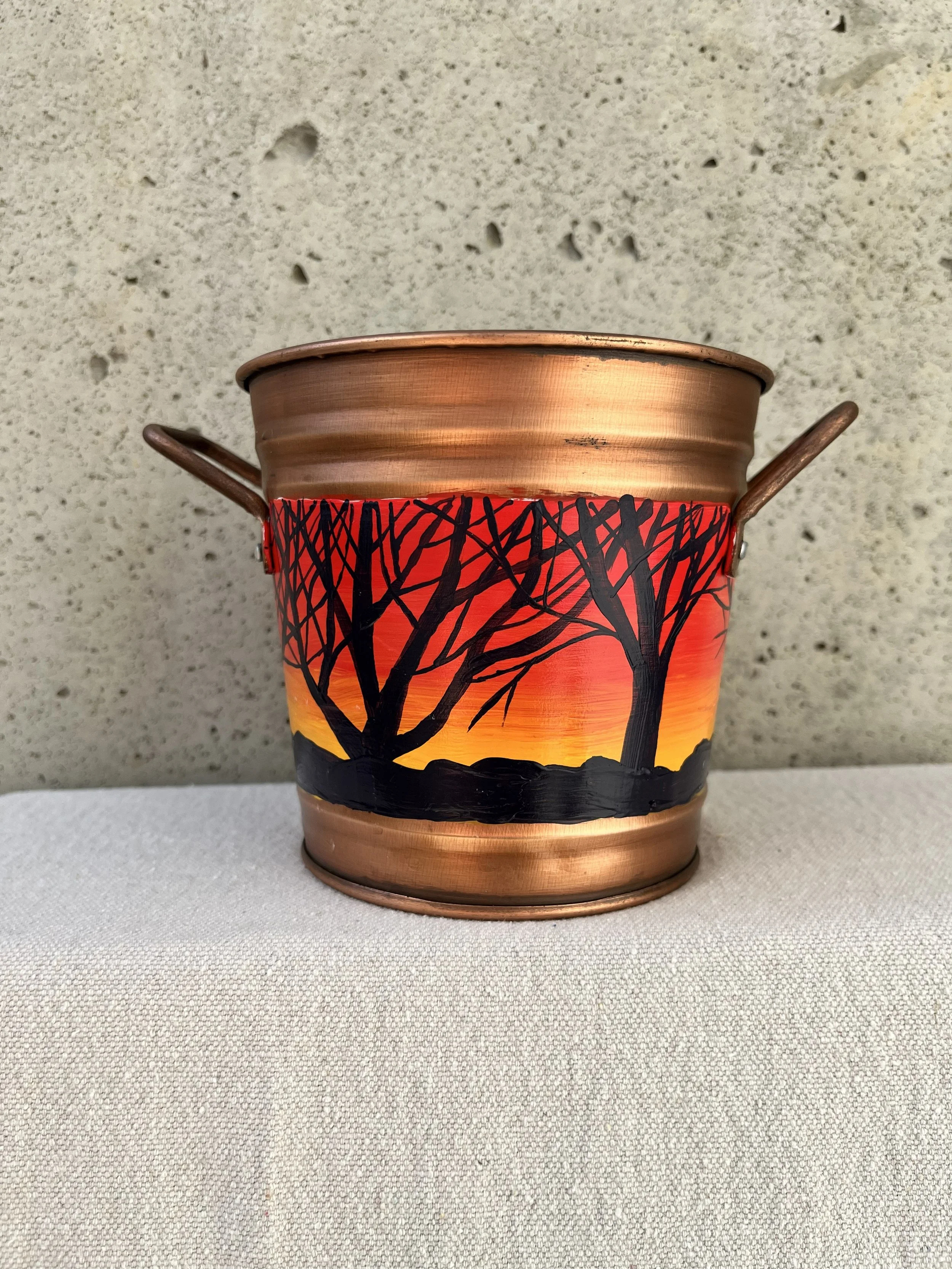 a handpainted landscape of bare branch trees silhouetted against a sunset. Painted on a copper color bucket.